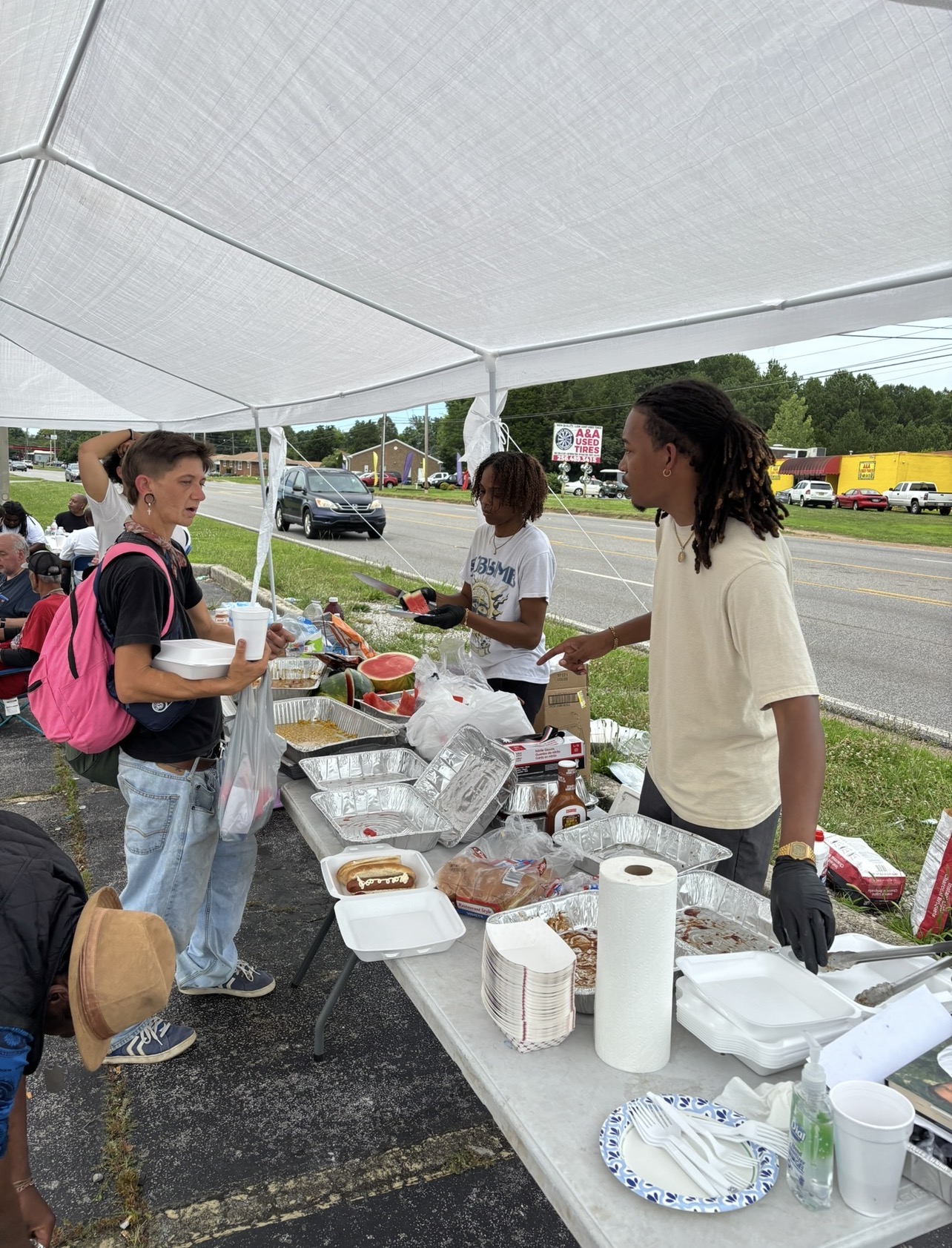 People gather at a table under a white canopy, serving food outdoors near a road with cars and trucks.