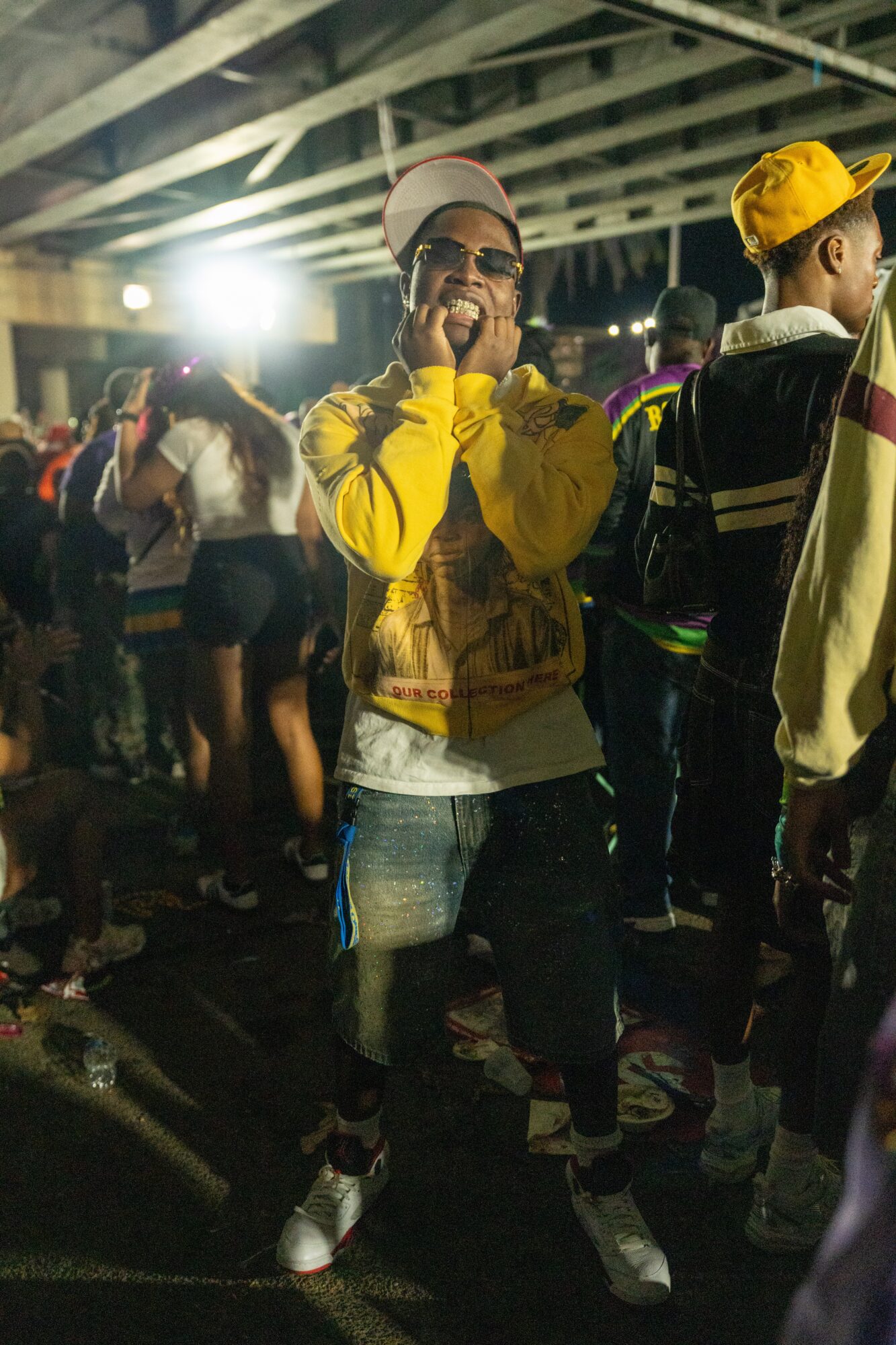 Person wearing a yellow jacket, white cap, and glasses, smiling and talking on a phone at a crowded event under a bridge.