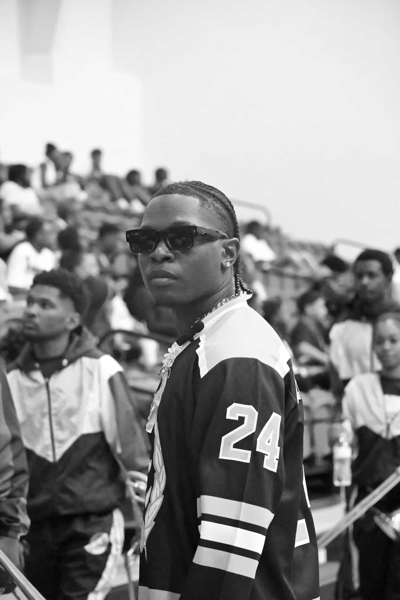 Young man wearing sunglasses and a sports jersey with the number 24, standing outdoors among a crowd.