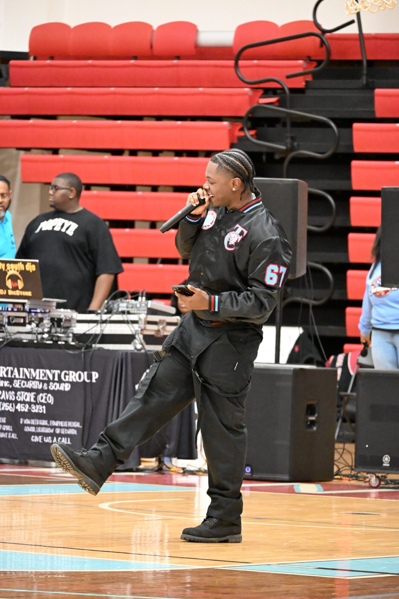 Person speaking into a microphone on a basketball court with red bleachers in the background.