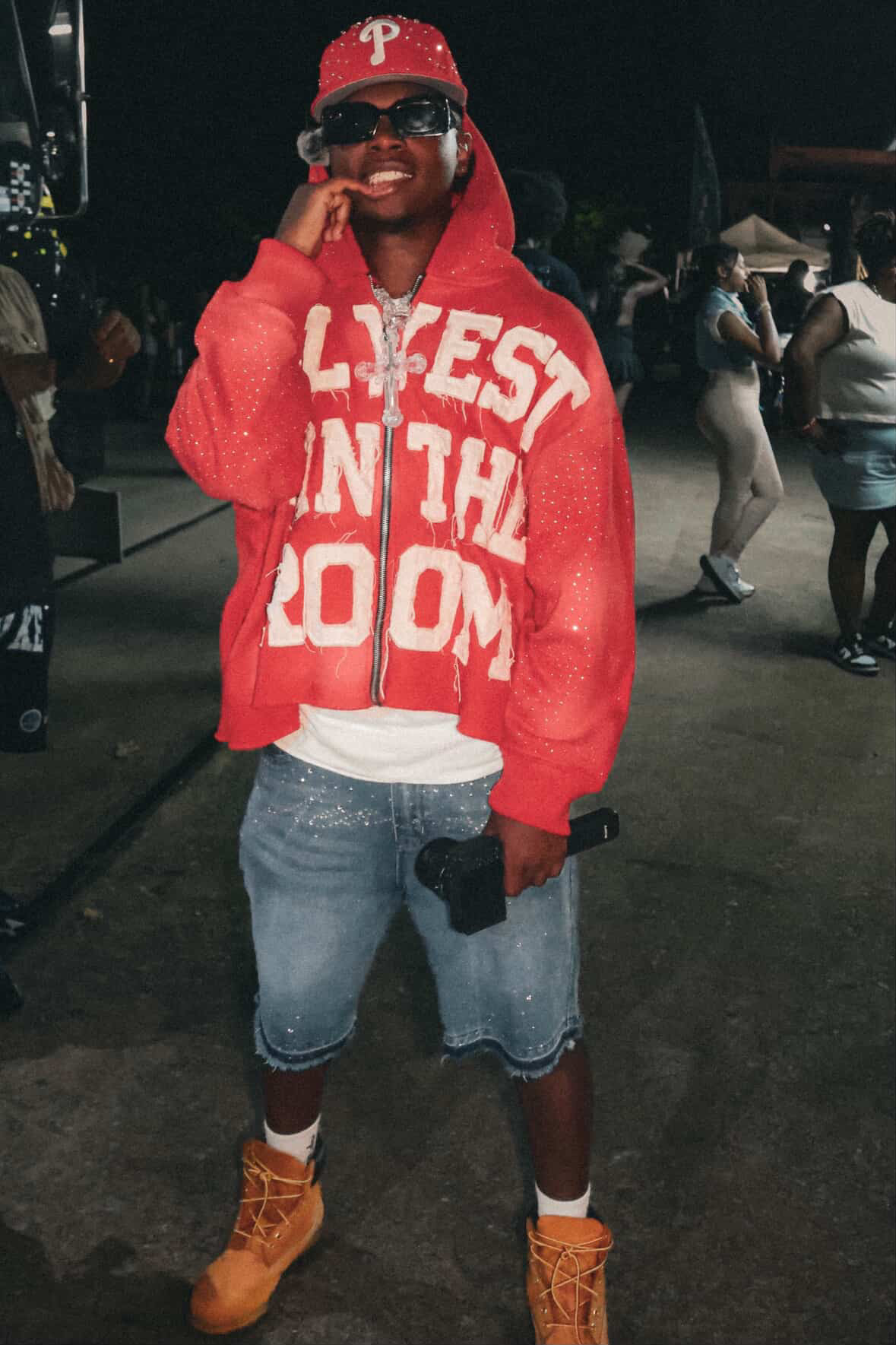 Person wearing a red hoodie, denim shorts, orange boots, and a red cap, smiling at night.