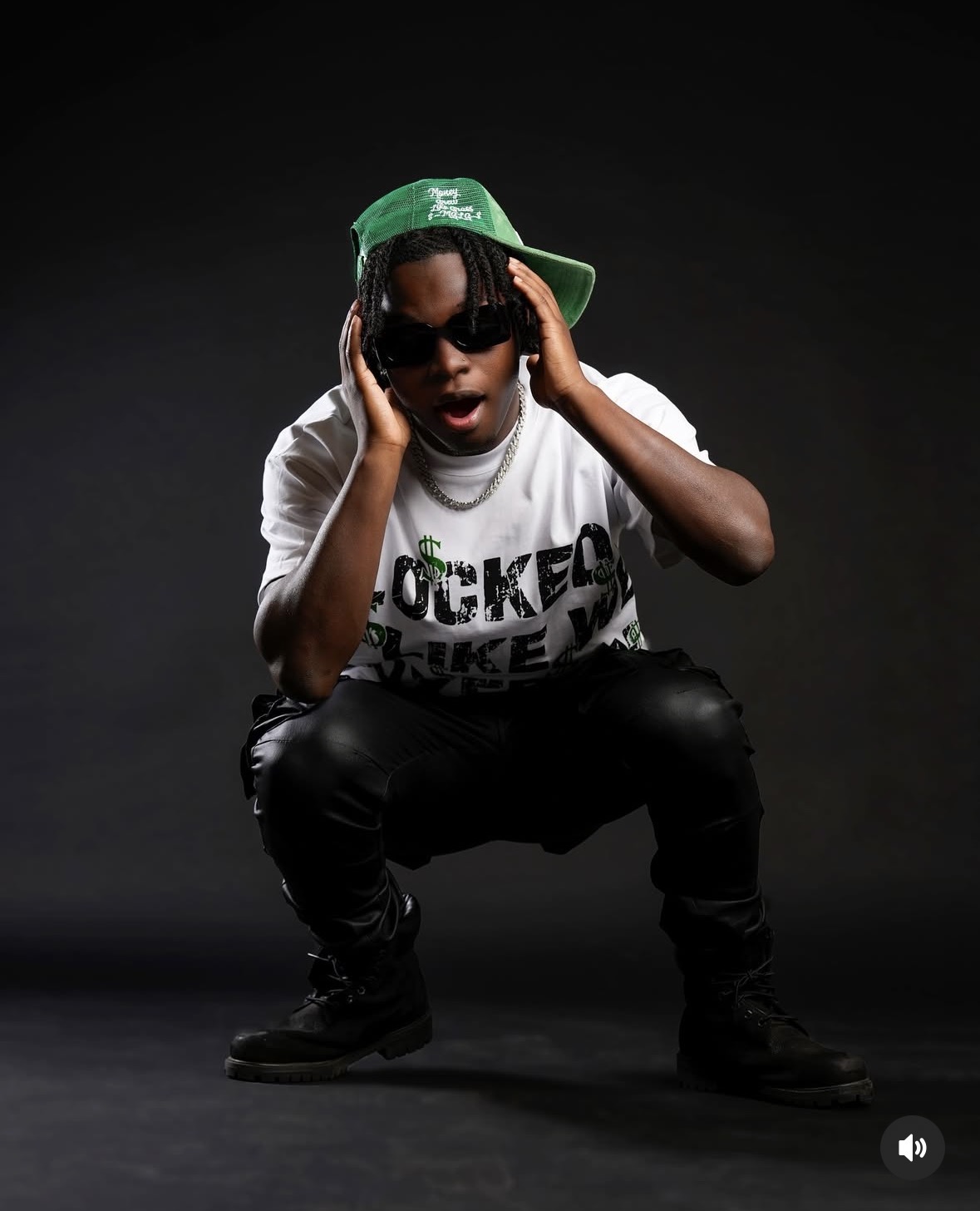 Person crouching, wearing sunglasses, a green cap, and a white T-shirt, against a dark background.