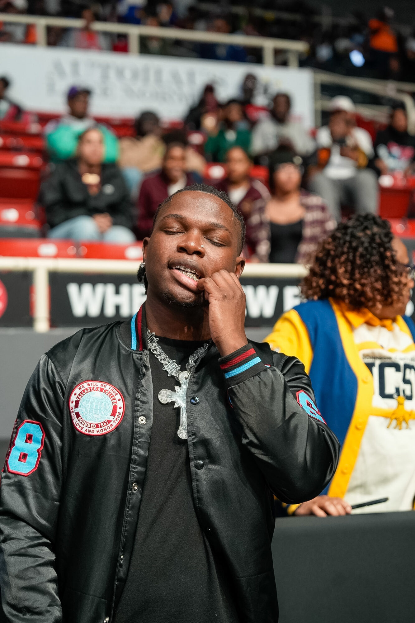 Person with eyes closed, hand on face, in a black jacket, at a crowded outdoor event.