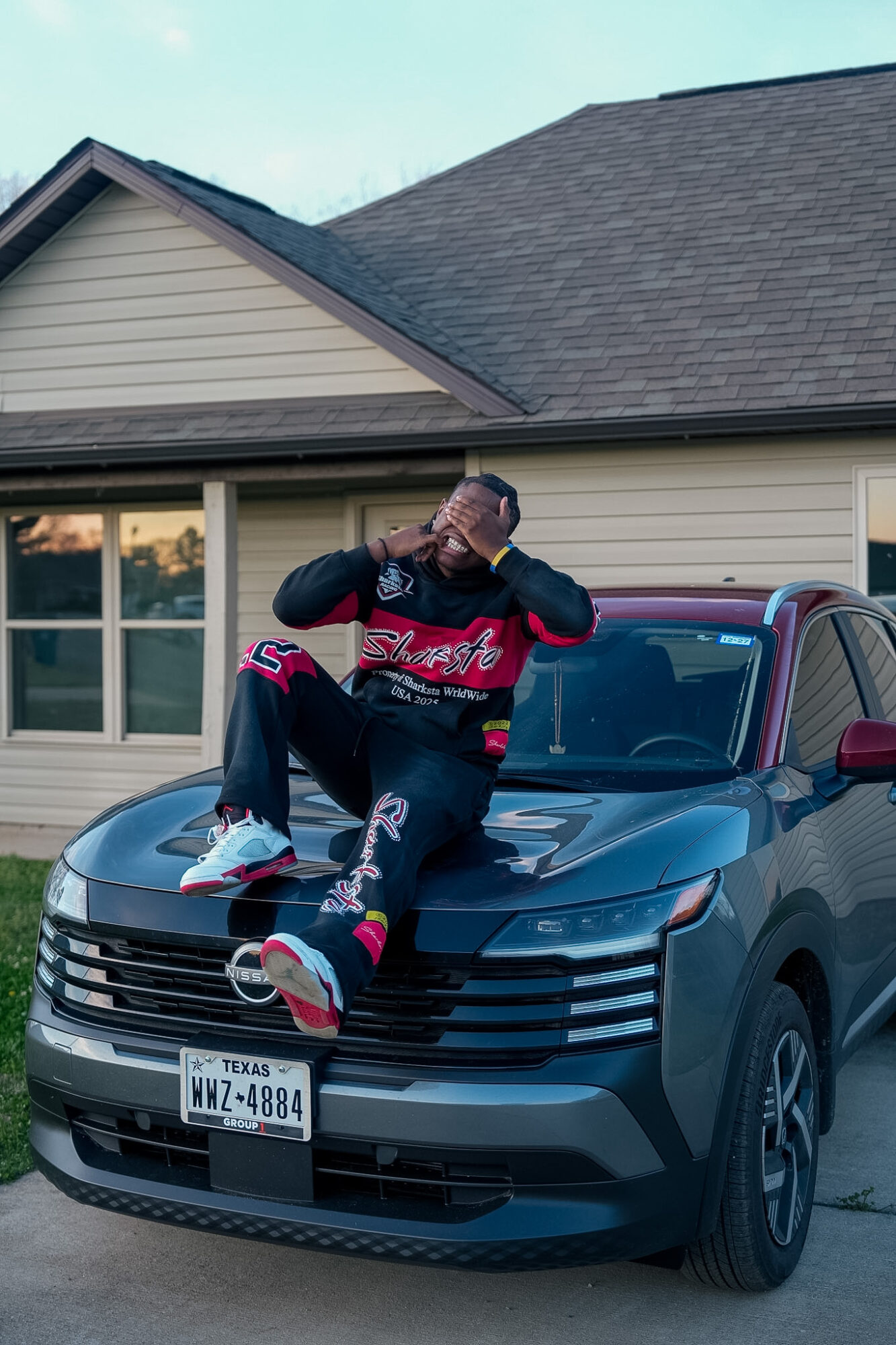 Person sitting on the hood of a black car, wearing a red and black jacket, glasses, and sneakers, in front of a house.