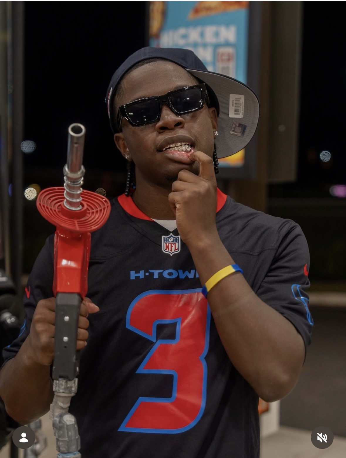 Person wearing sunglasses, a cap backward, and a sports jersey, holding a toy lightsaber, with a fast-food sign in background.