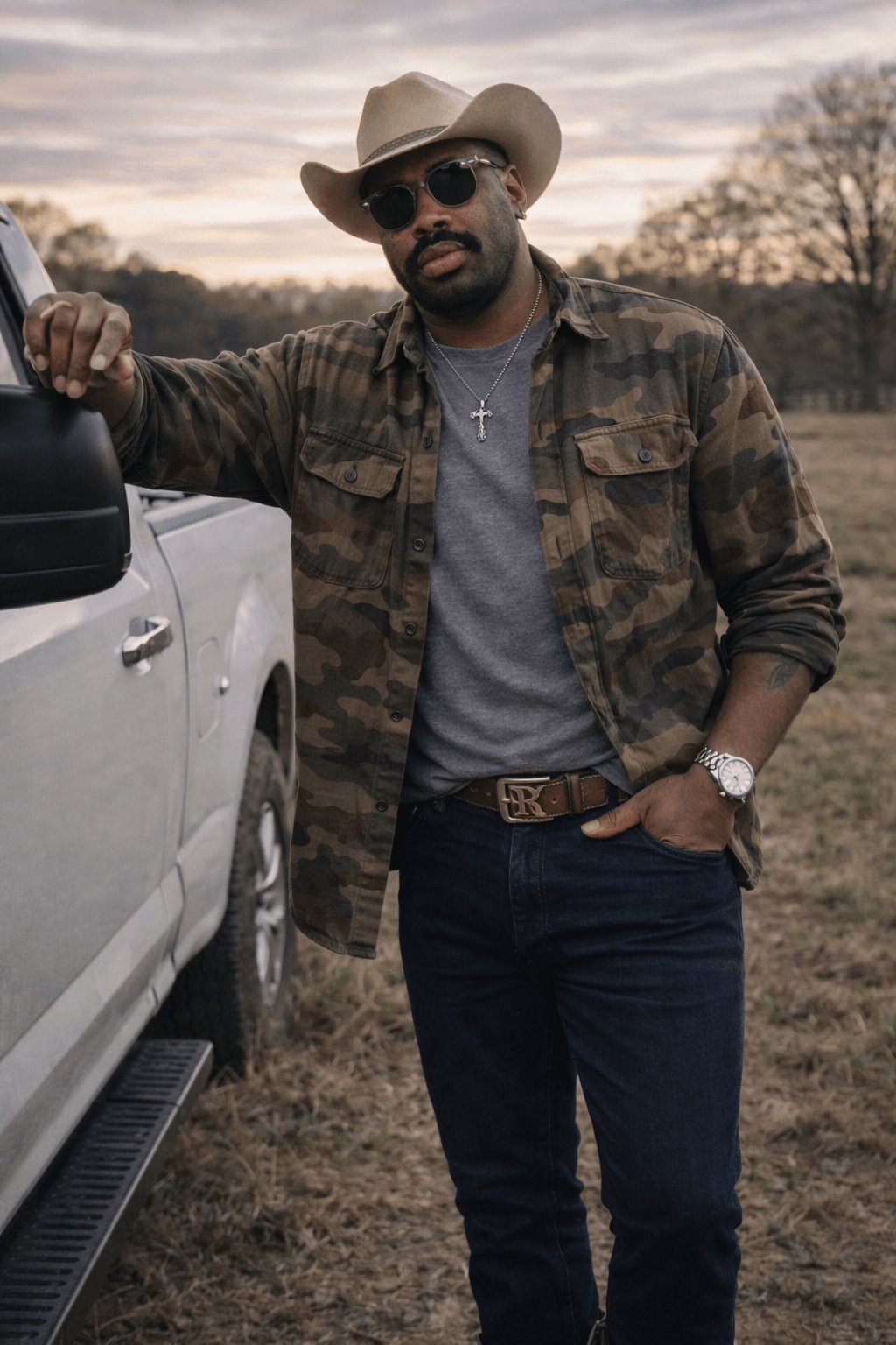 Man wearing sunglasses, hat, camouflage jacket, and cross necklace standing outdoors next to a white vehicle.