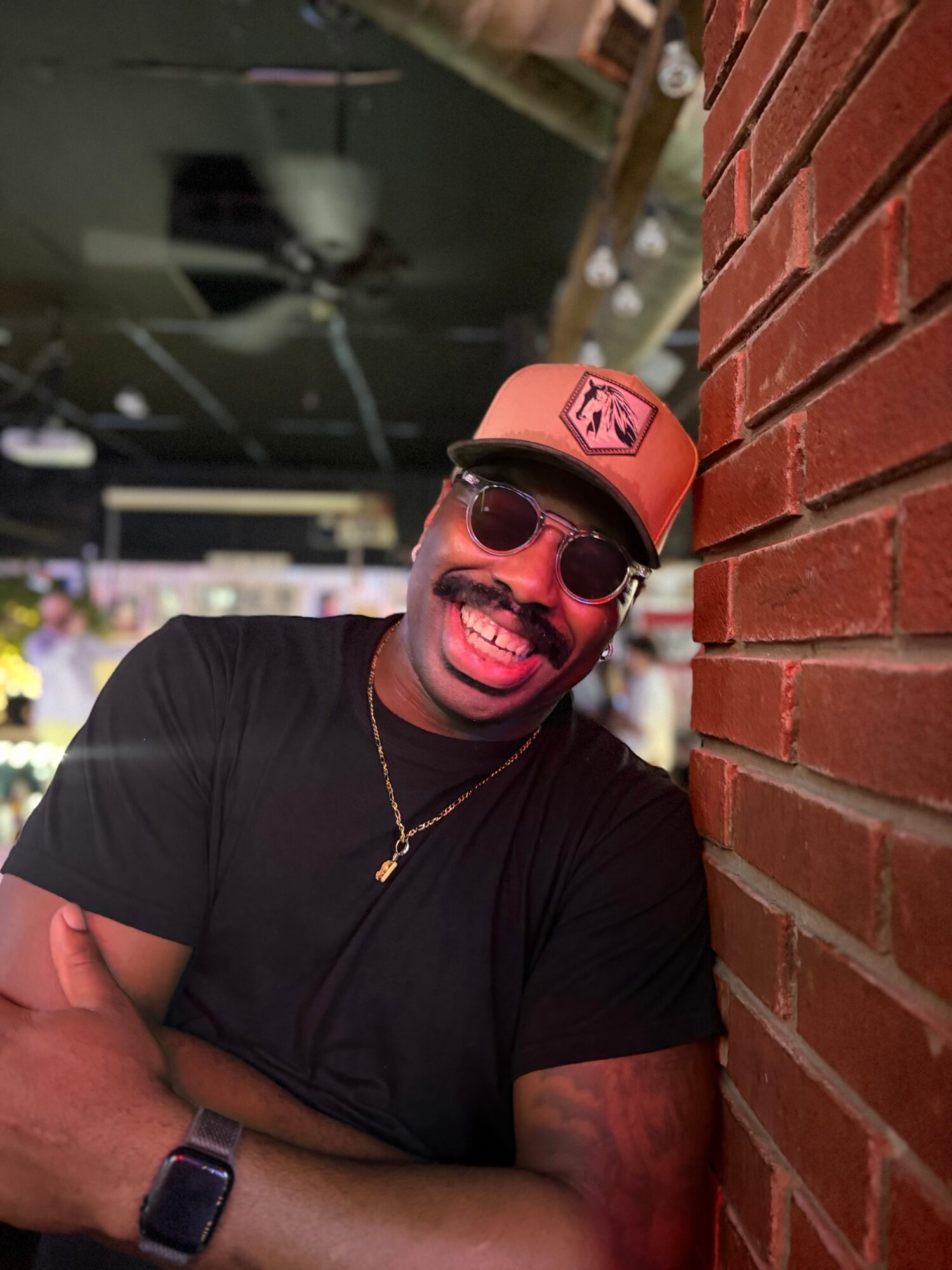Man wearing sunglasses and a cap, smiling, leaning against a brick wall indoors.