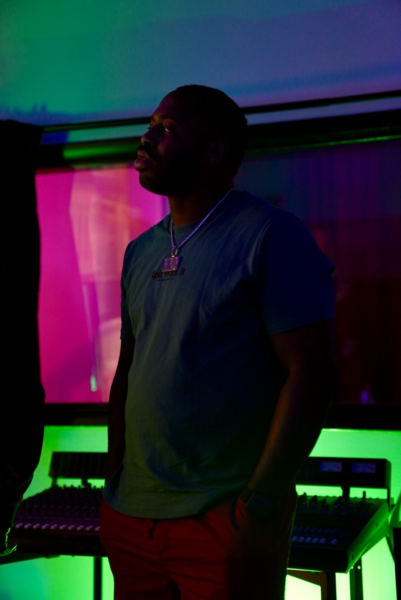 Person standing in a dark room with colorful lighting, wearing a t-shirt and necklace, with electronic equipment in the background.