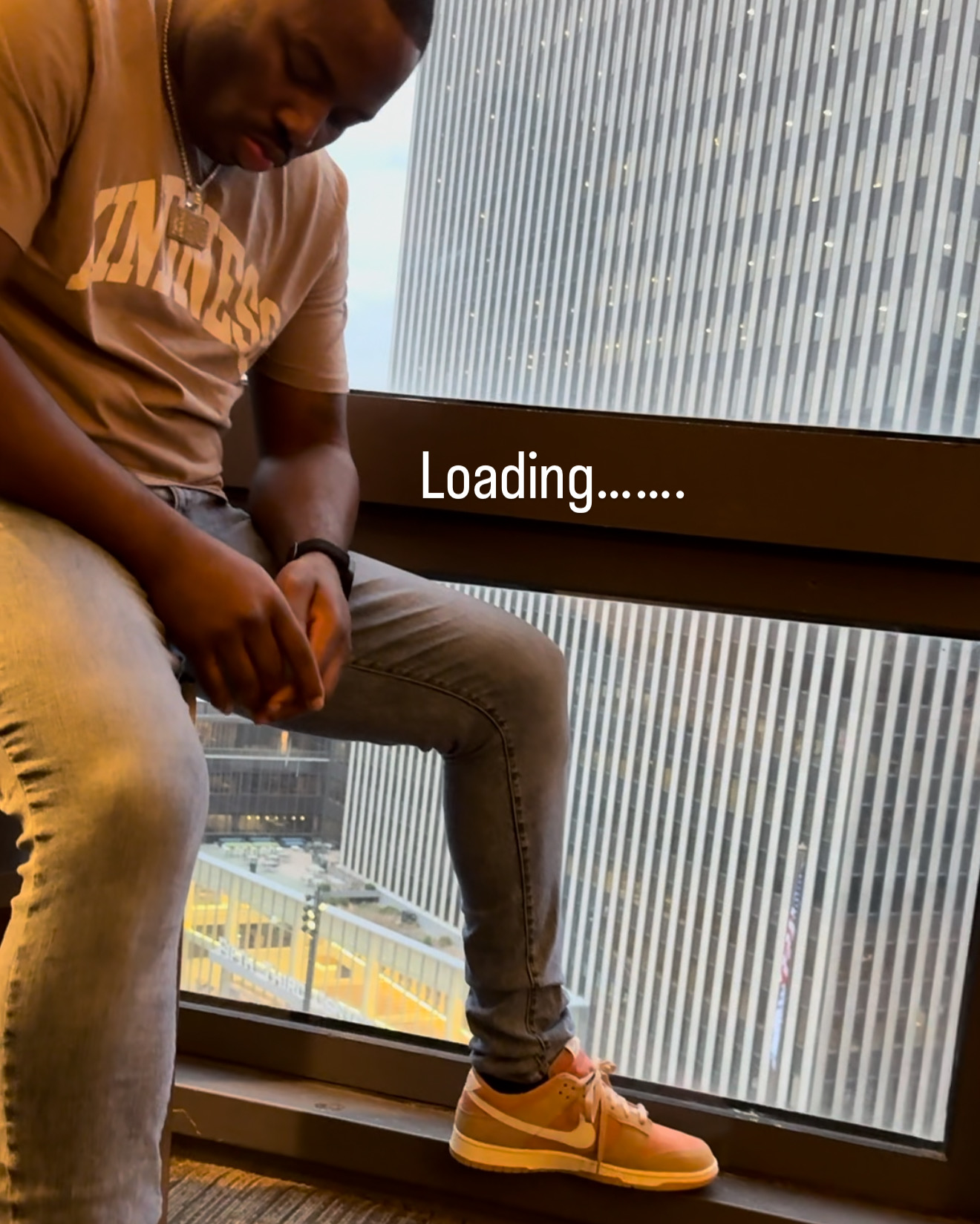 Person sitting by window with city view, loading screen overlay, wearing beige pants and yellow sneakers.