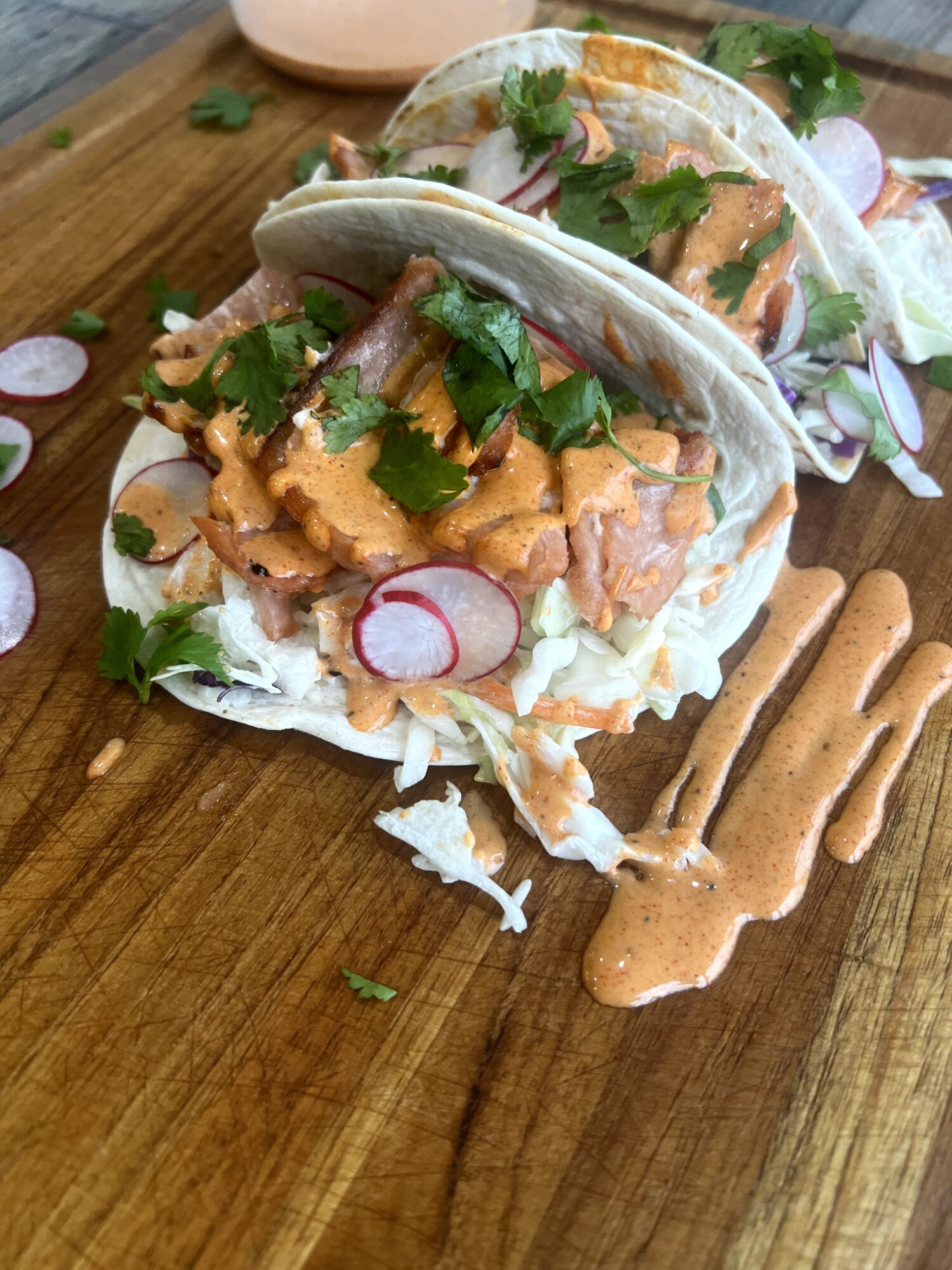 Two tacos filled with meat, cilantro, radish slices, and sauce on a wooden surface with extra sauce nearby.