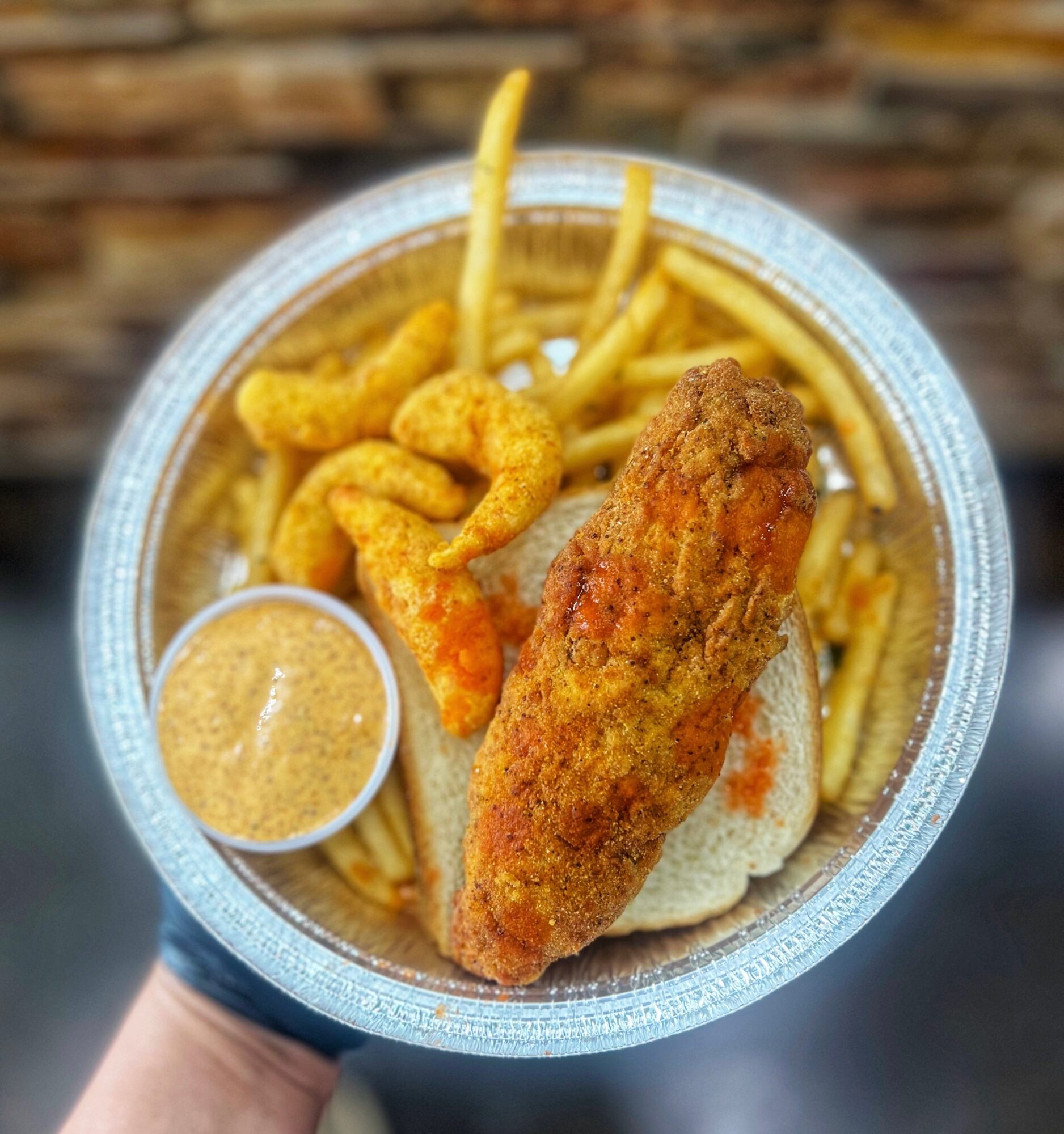 Container with fried fish, French fries, shrimp, and dipping sauce, held by a hand against a brick wall background.