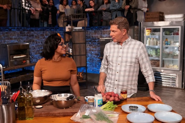 Two people talking in a kitchen with plates, bowls, and ingredients on the counter, and a group of people in the background.
