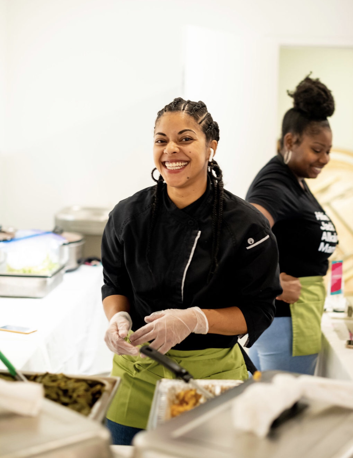 Two women working in a kitchen, smiling, wearing black shirts and green aprons, with food and kitchen tools around them.