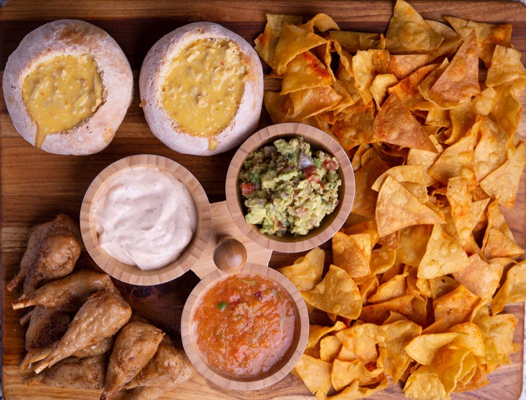 Baked eggs in bread bowls, chips, guacamole, salsa, sour cream, and sausage on a wooden surface.