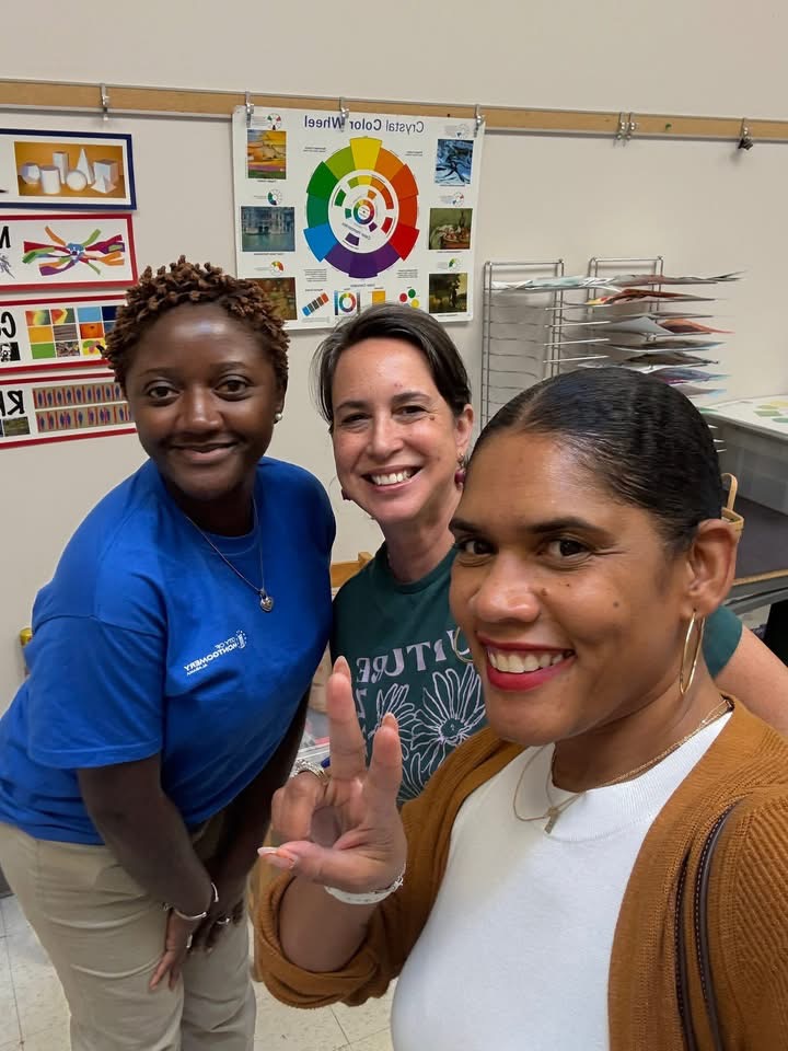 Three women smiling and taking a selfie in a classroom with educational posters and supplies behind them.