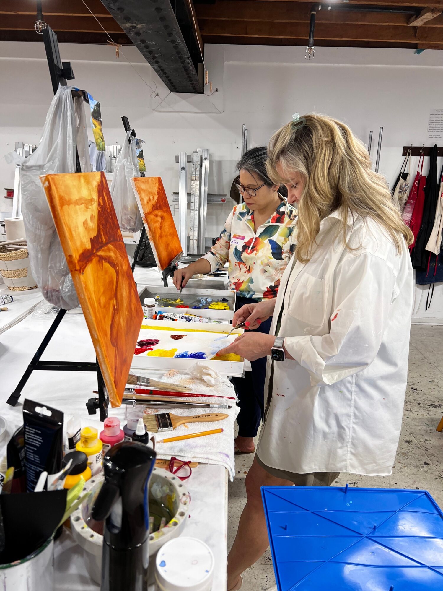 Woman with blonde hair in a white coat working on a colorful art project at a cluttered table.