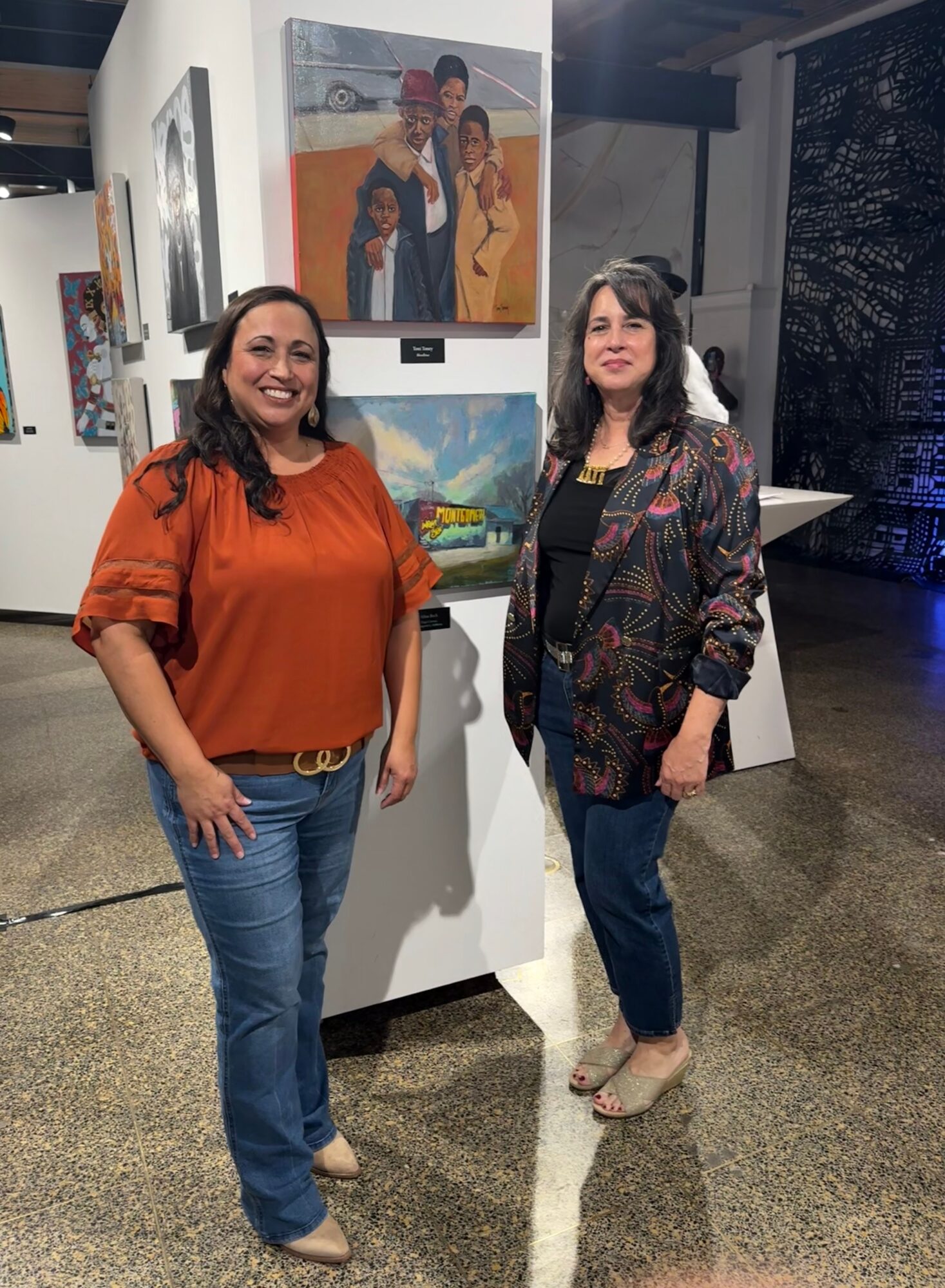 Two women stand in an art gallery, smiling, with paintings displayed behind them.