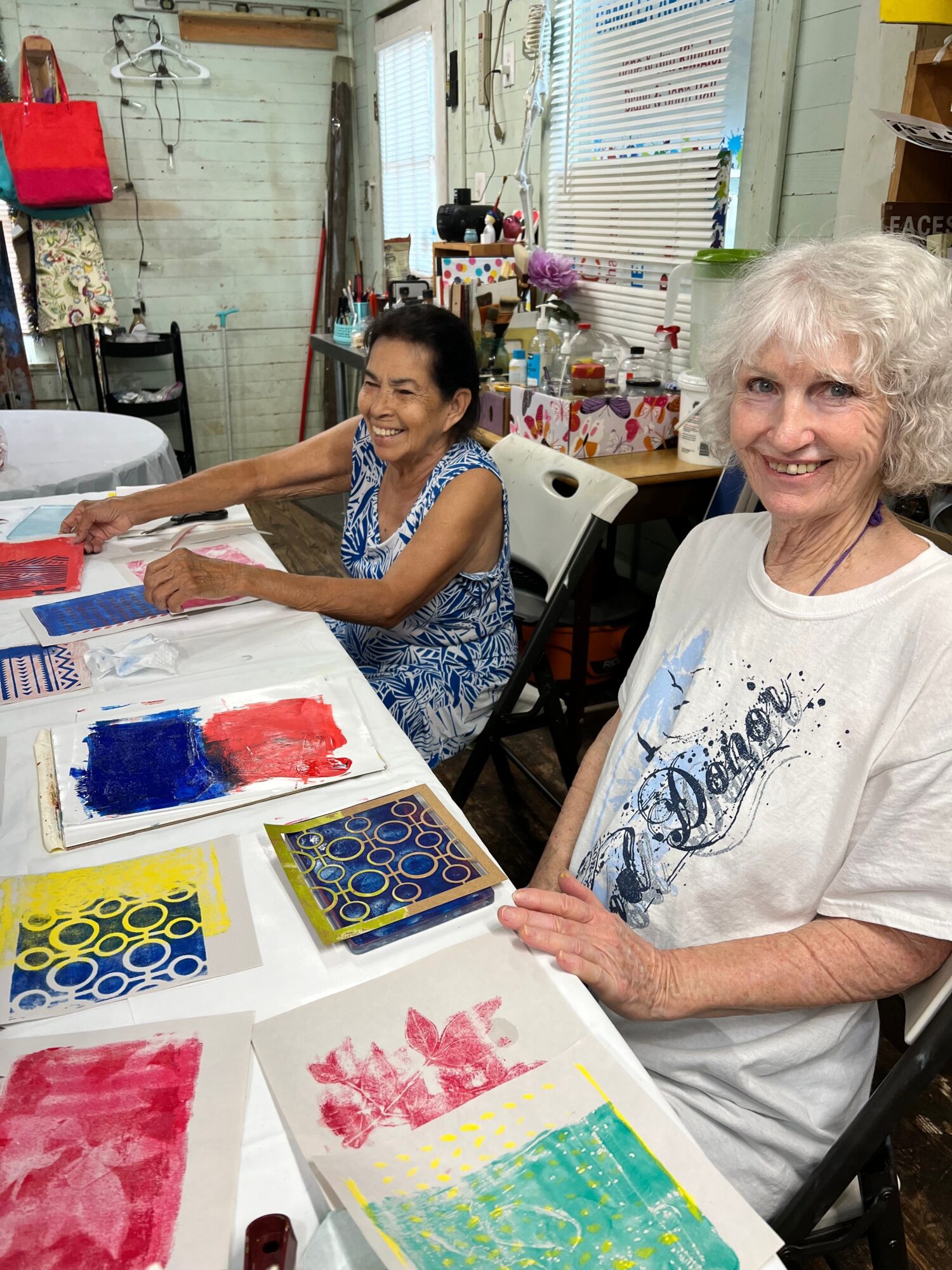 Two women sitting at a table with colorful paintings and art supplies, smiling and engaging in art activity.