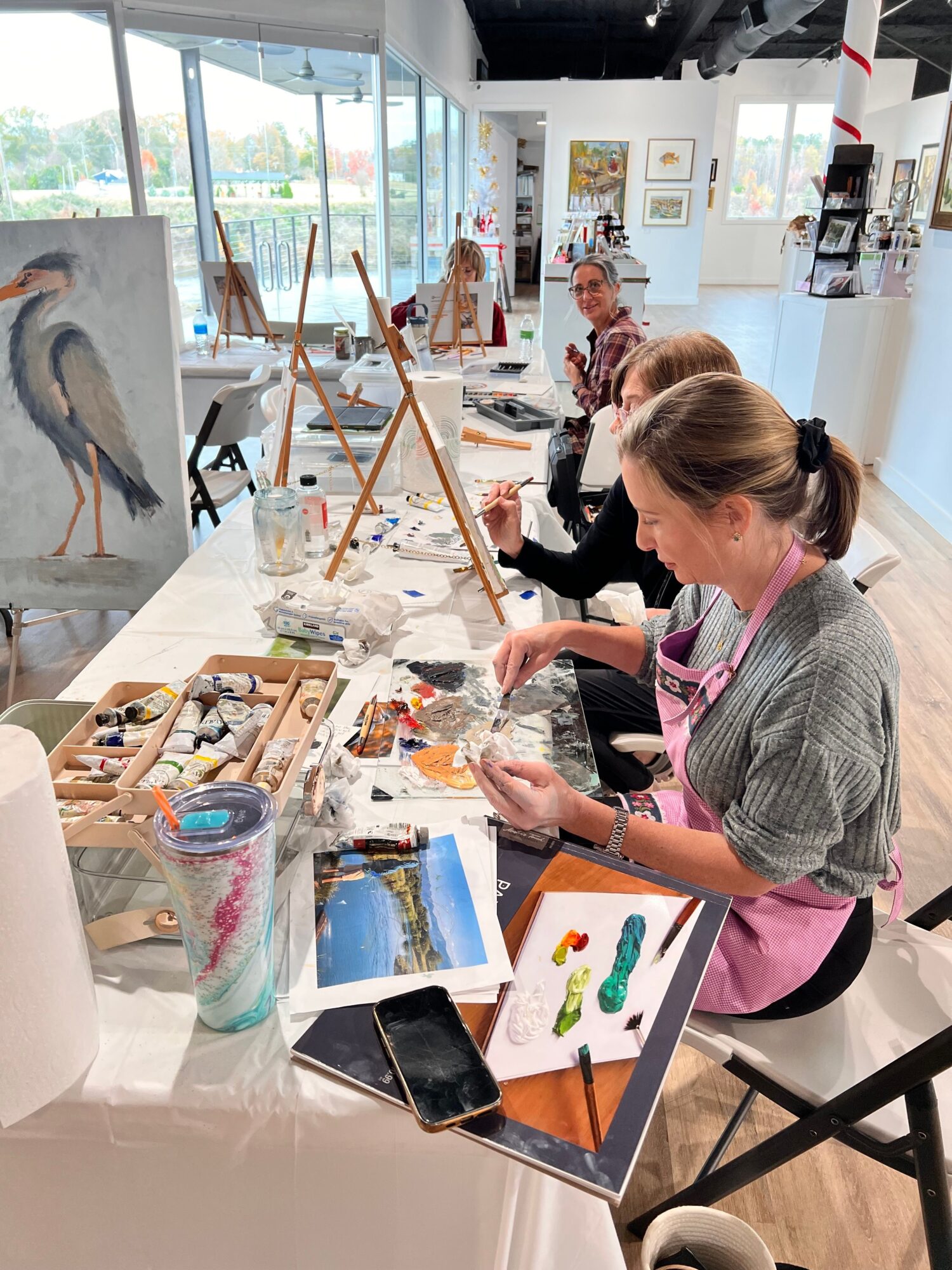 People working on art projects at a table with paints, brushes, and reference images in a bright room.