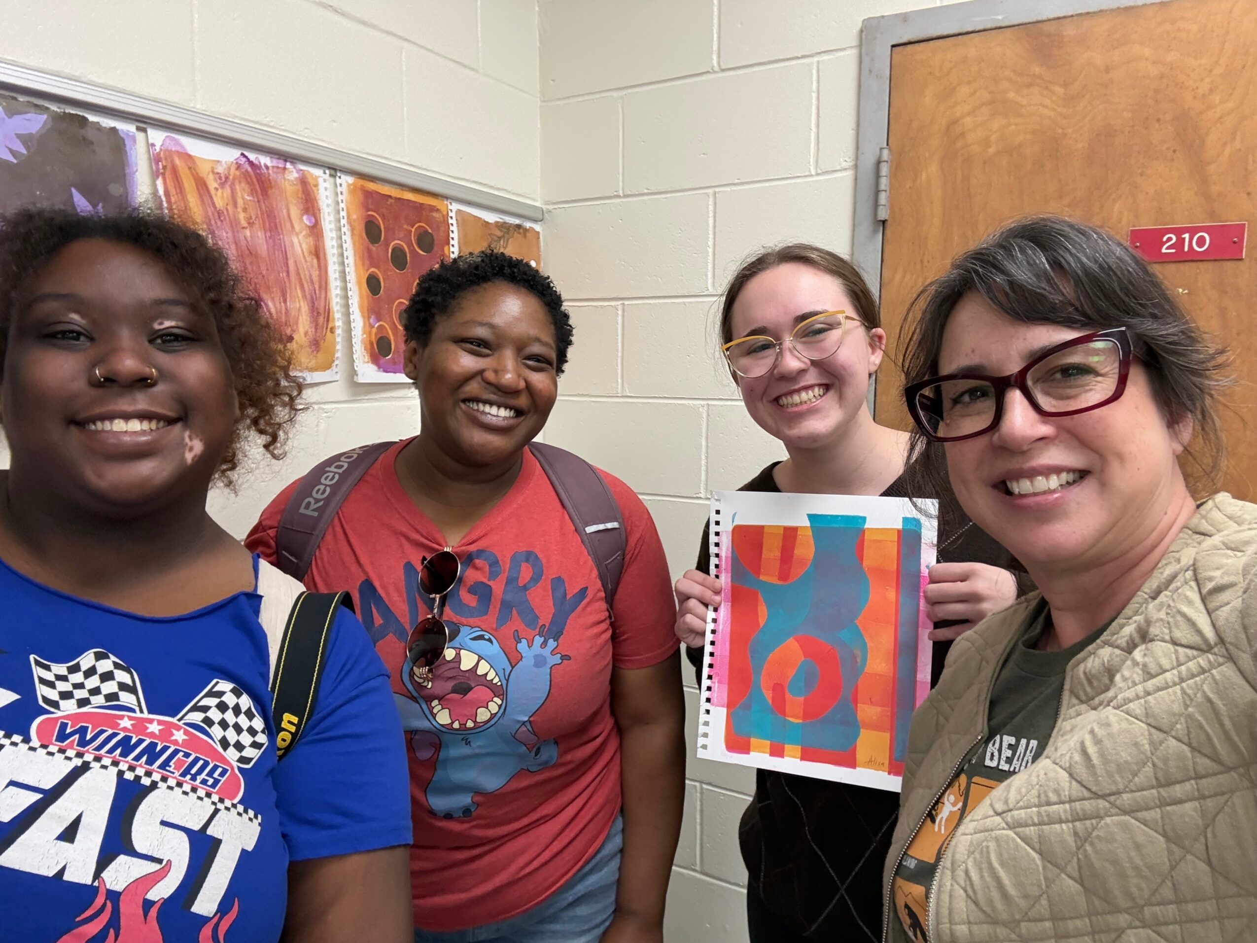 Four women smiling indoors, holding colorful artwork, standing in front of a wall with artwork and a door with a pink label.