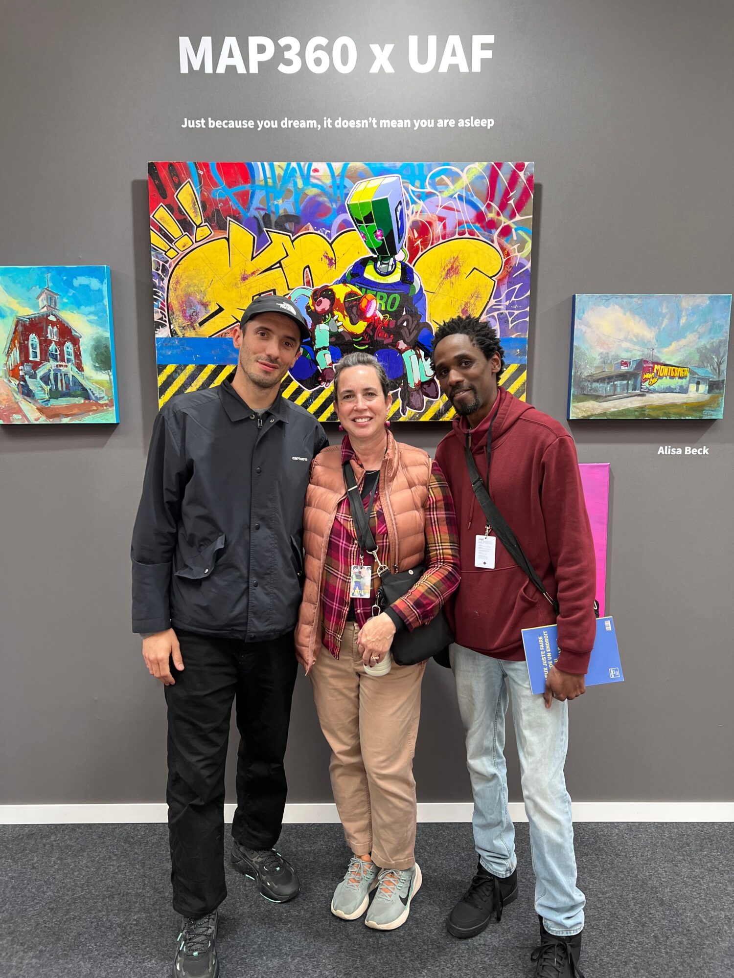 Three people standing in front of colorful graffiti art, holding books, in an indoor setting.