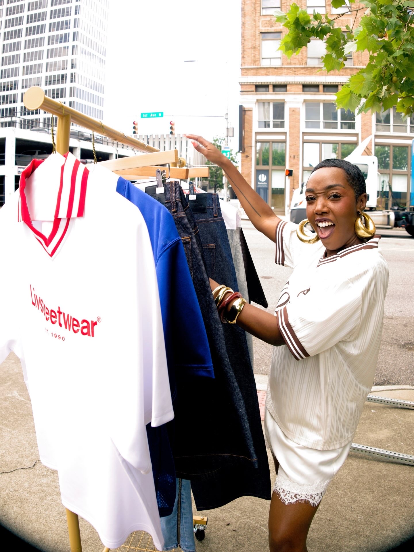 Woman smiling and shopping outdoors with clothing on a rack, city buildings in background.