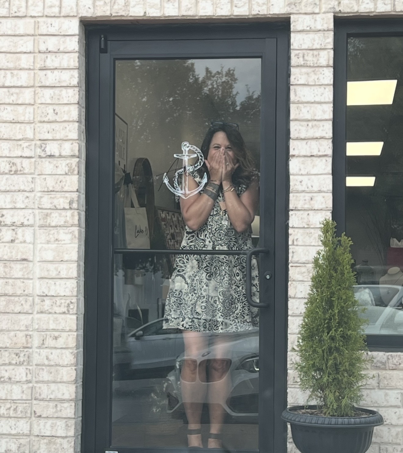 Woman taking a photo through a glass door, reflection shows her holding a phone, brick wall background, potted plant nearby.