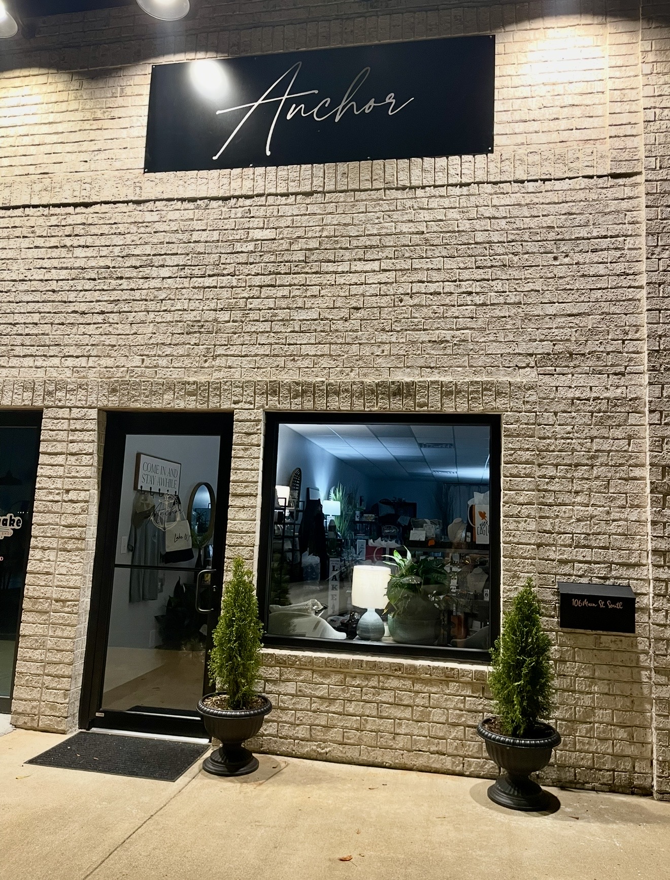 Storefront with a sign reading 'Anchor' above the entrance, two potted plants outside, and a large window showing interior lighting.