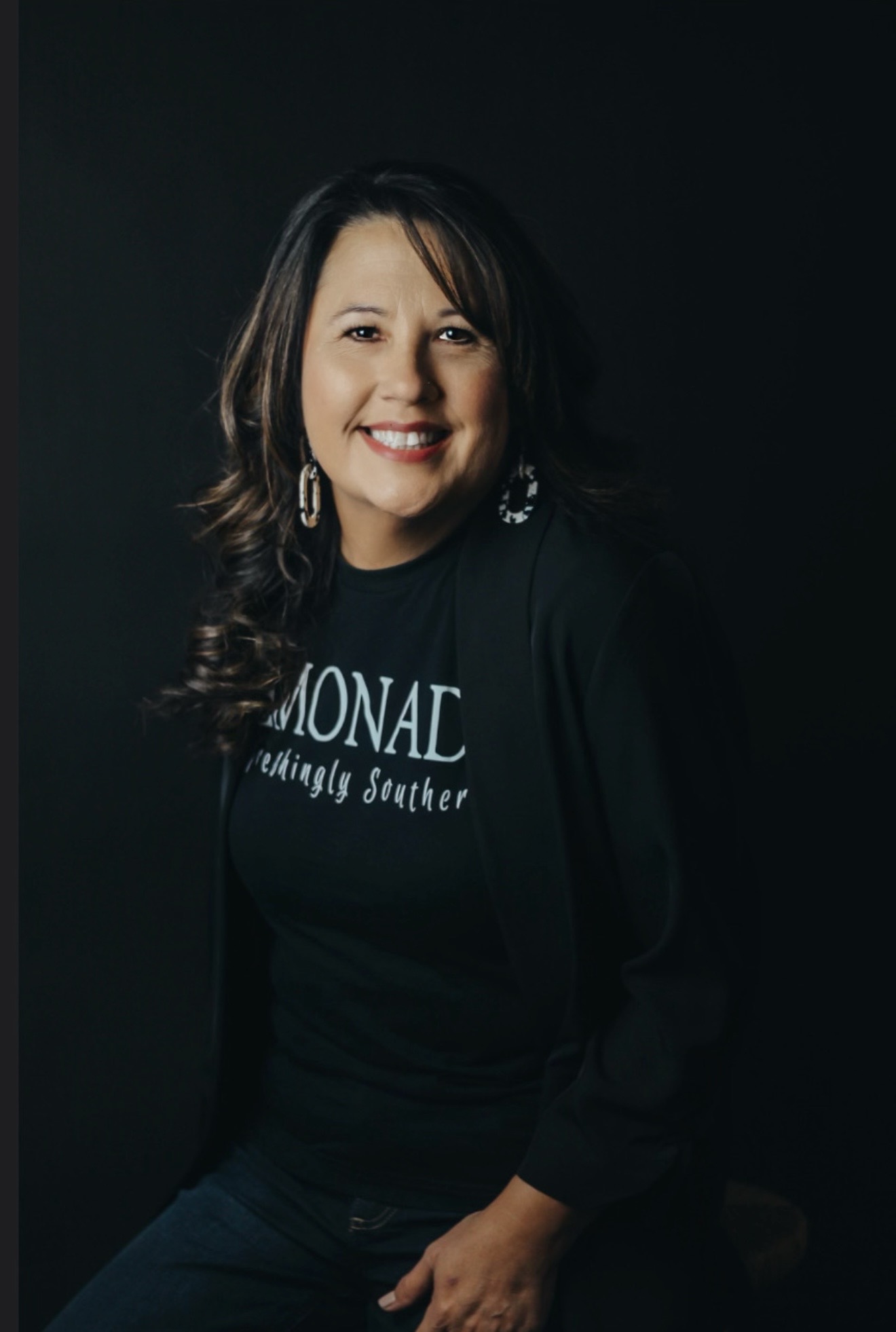 Woman with long dark hair smiling, wearing a black top with text, earrings, and a dark blazer, against a dark background.