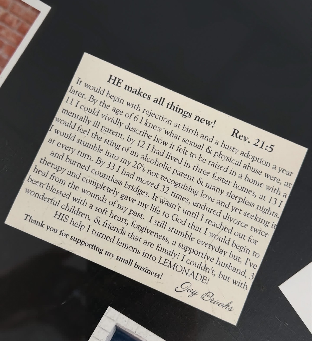 Close-up of a printed prayer card with a Bible verse, titled 'HE makes all things new! Rev. 21:5', on a dark surface.