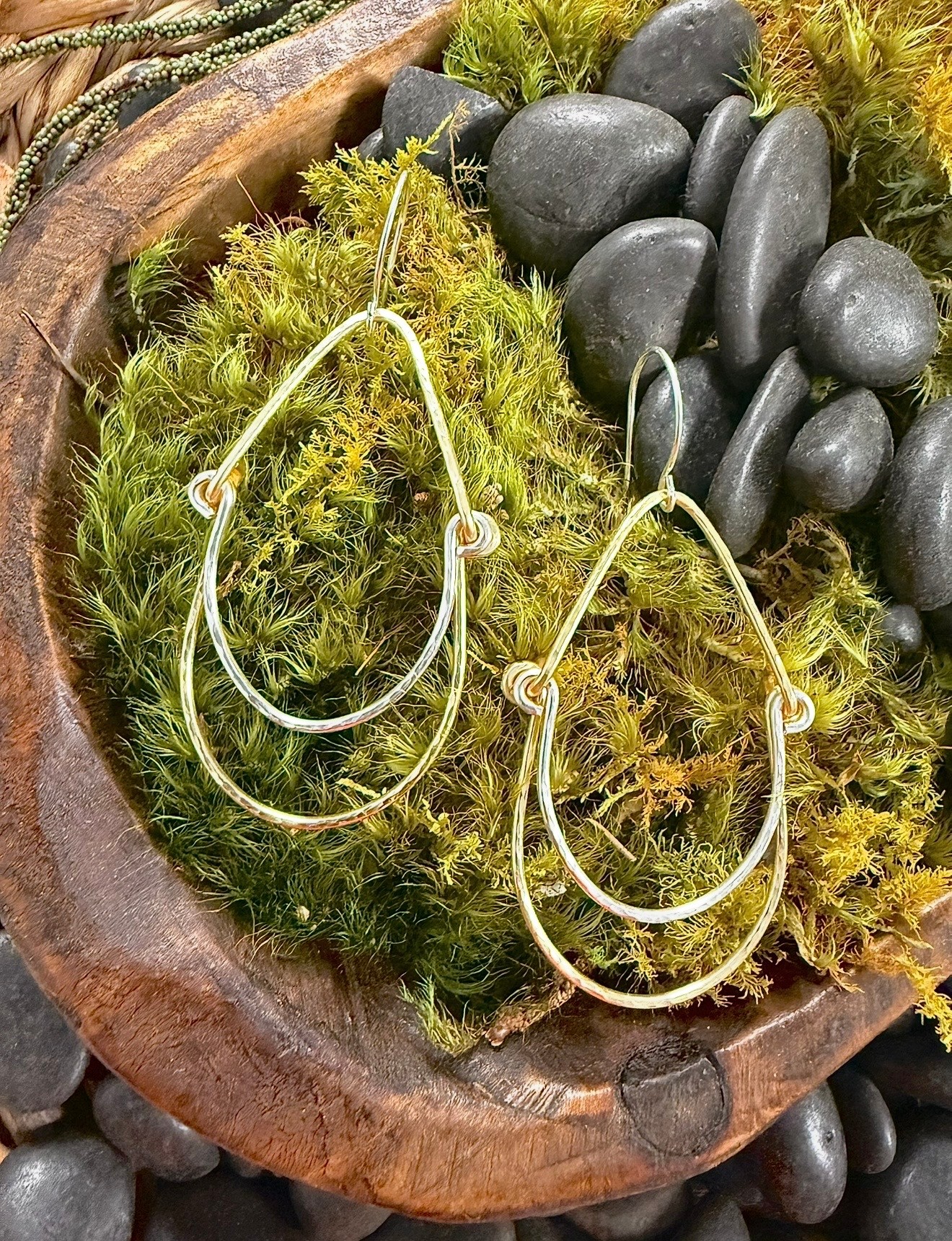 Pair of gold hoop earrings with decorative knots, placed on green moss with black stones in the background.