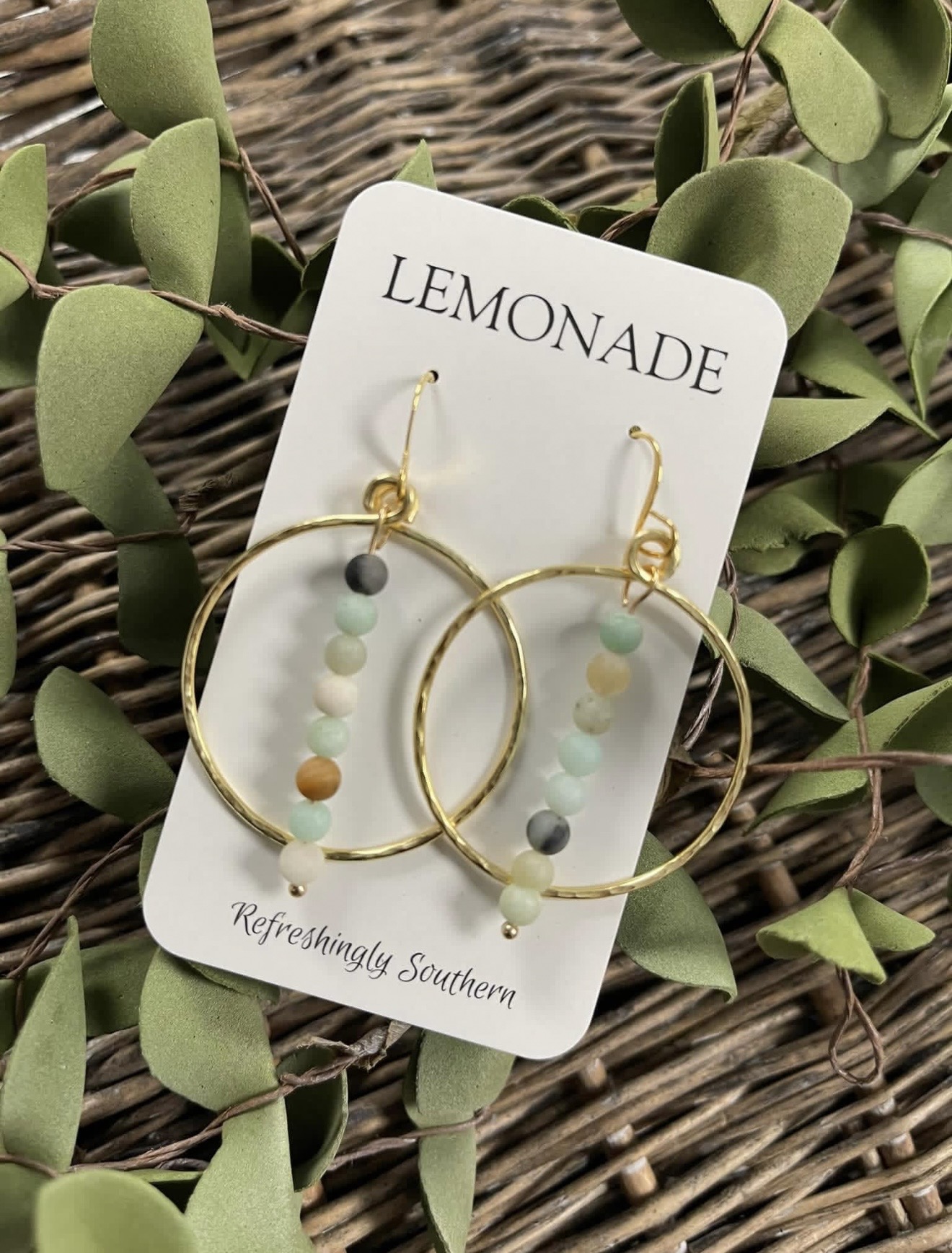 Pair of gold hoop earrings with pastel beads on a white card labeled 'LEMONADE' surrounded by green leaves and woven background.