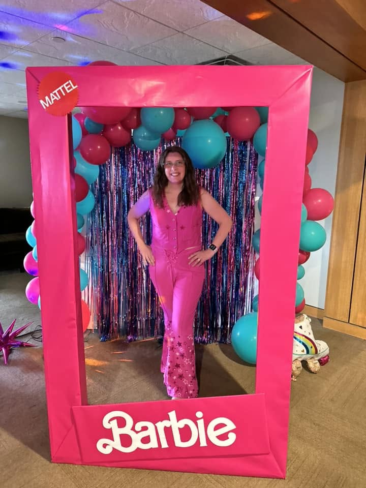 Woman in pink jumpsuit inside a Barbie-themed photo frame with balloons and a colorful backdrop.
