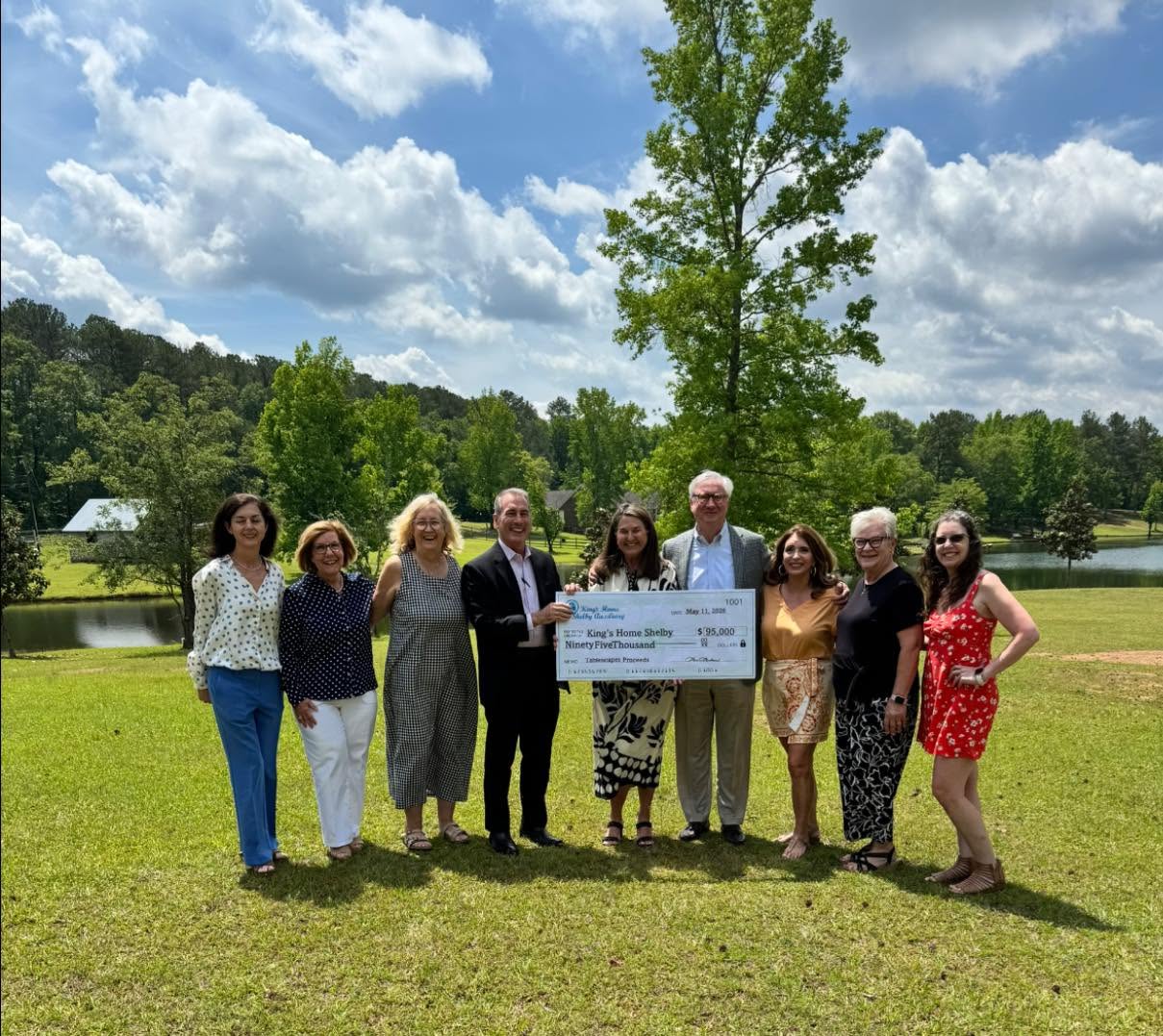Group of nine people standing outdoors on grass, holding a large check, with trees and a lake in the background.
