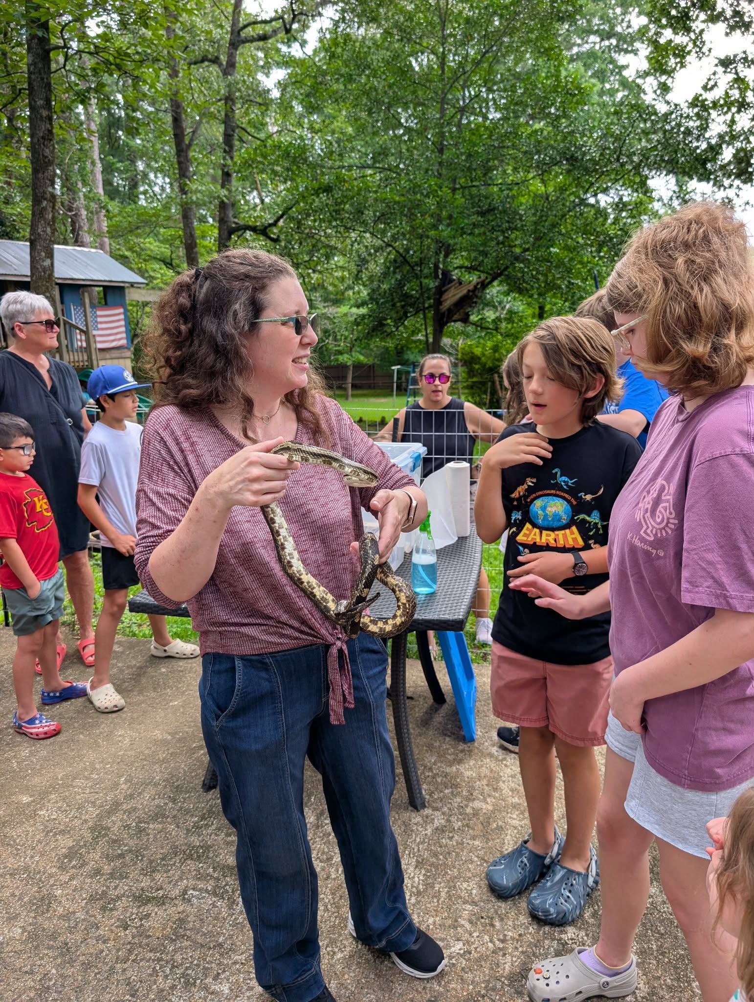 Group of children and adults outdoors in a wooded area, with a woman showing a snake to a boy, others watching.