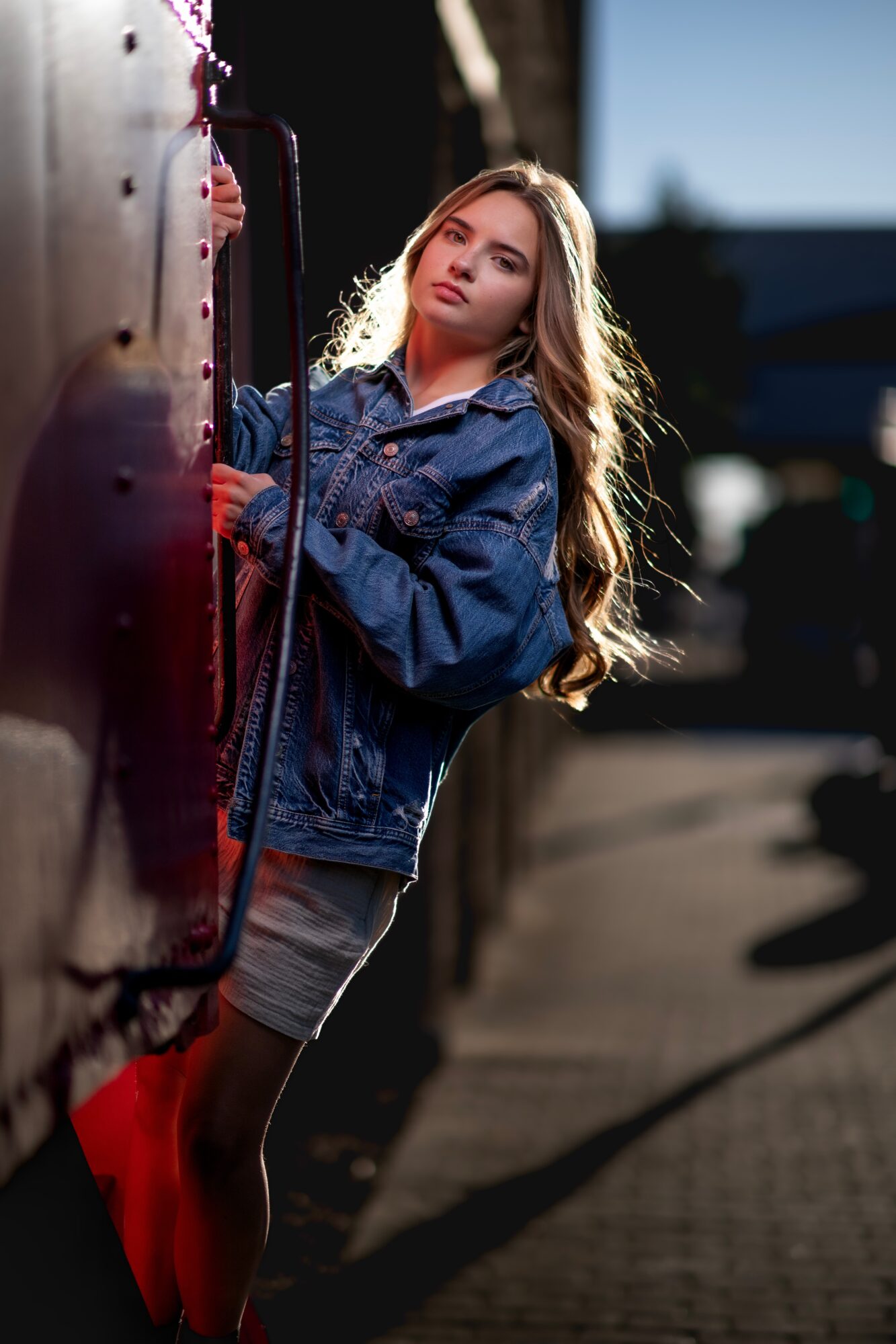 Young girl with long hair in a denim jacket leaning against a red train, sunlight highlighting her face and hair.