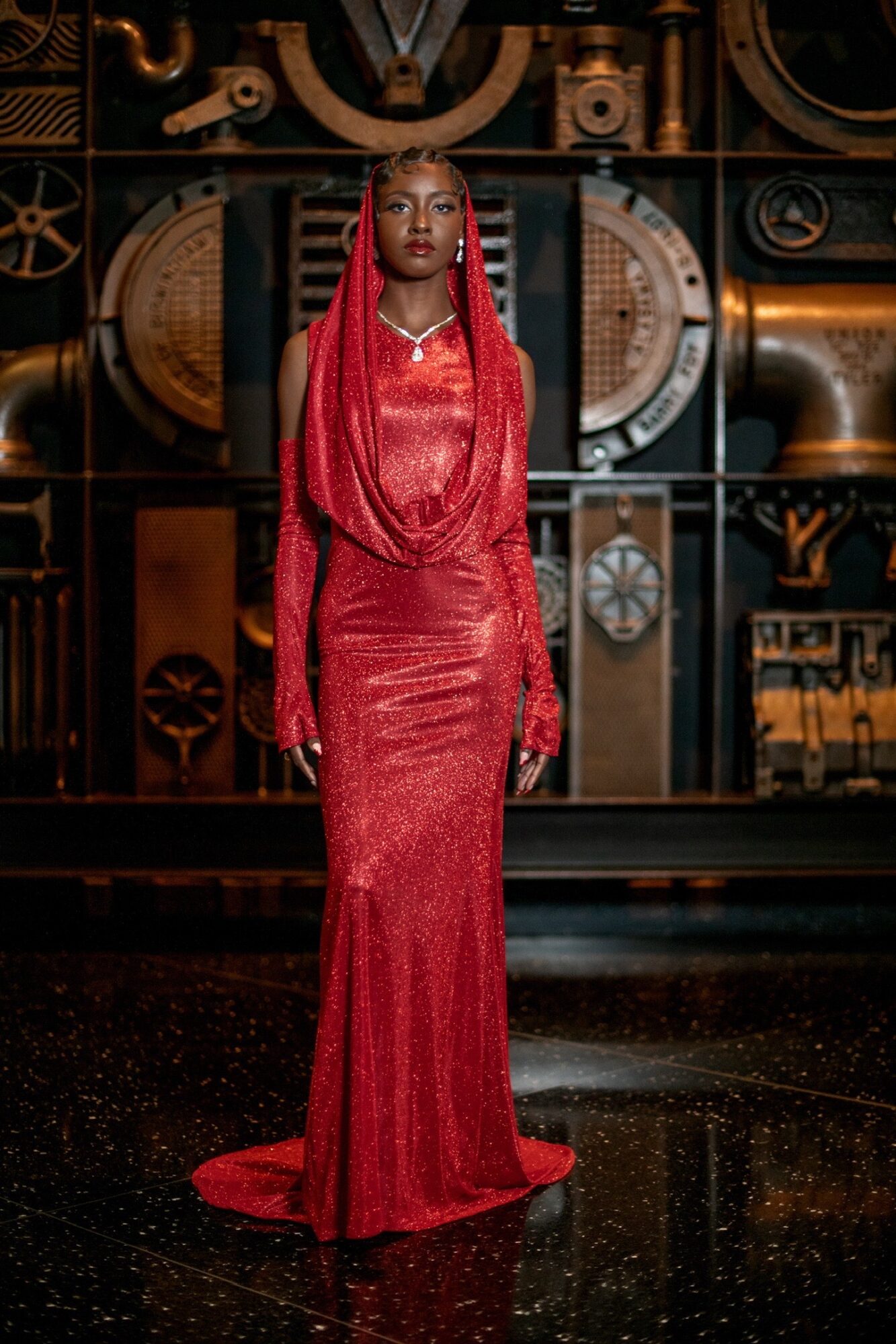 A woman in a red gown with long braids stands on a stage with mechanical and industrial background elements.