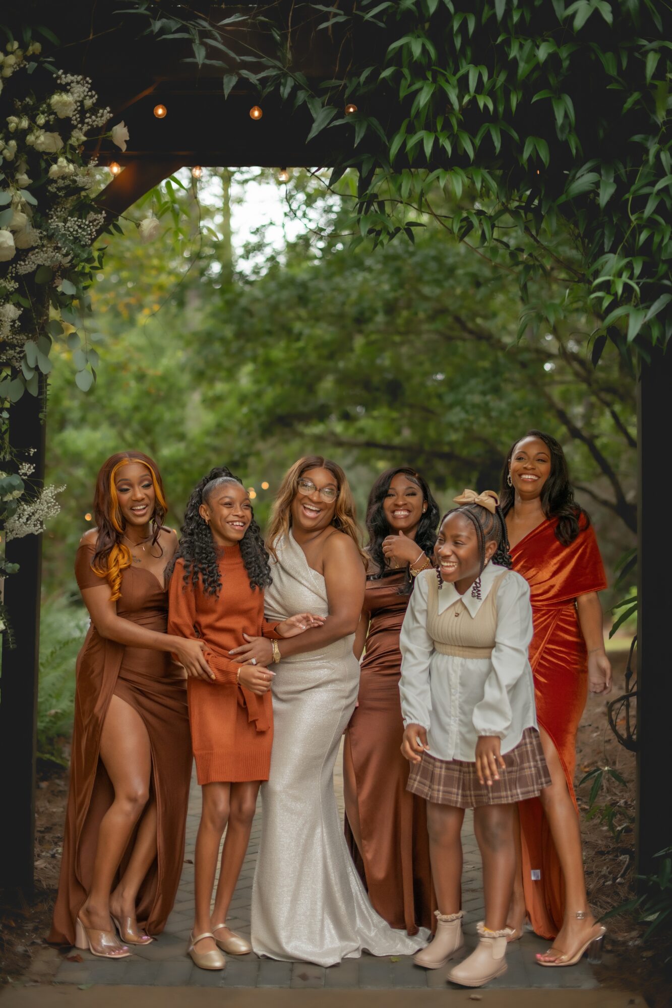 Group of six women standing outdoors under a tree, smiling, with greenery in the background.