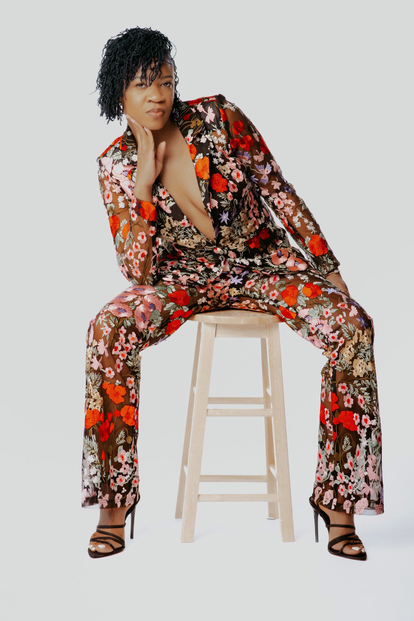 Woman with curly hair in a floral suit sitting on a stool, looking to the side, against a plain background.