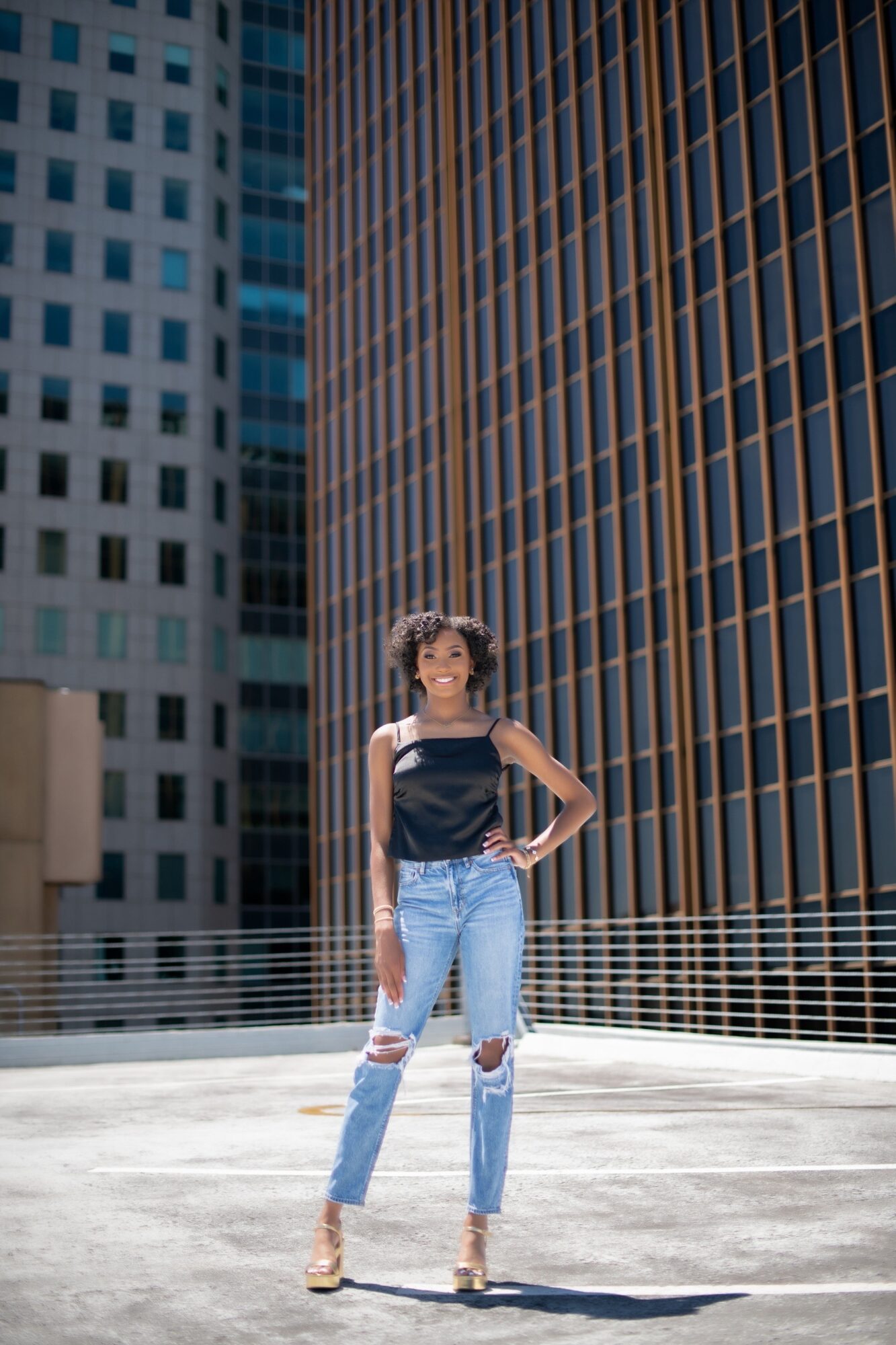 Young woman standing on rooftop with tall buildings in background, wearing a black top and ripped jeans.