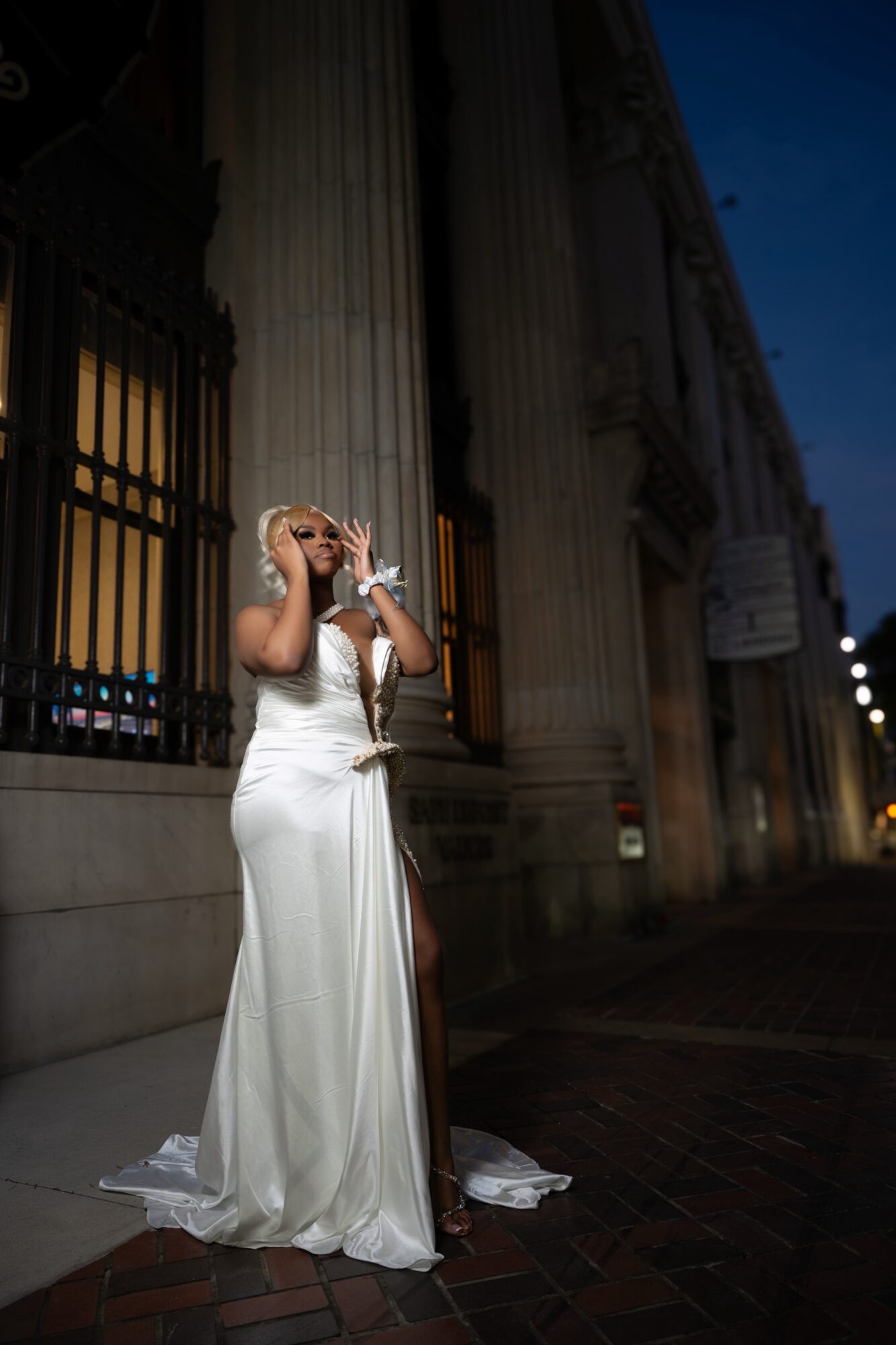 Woman in a white dress standing outside at night, holding a camera, with a building in the background.