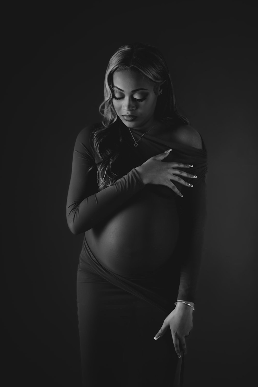 Pregnant woman with long hair, wearing a long dress, gently touching her belly, in a dark studio setting.