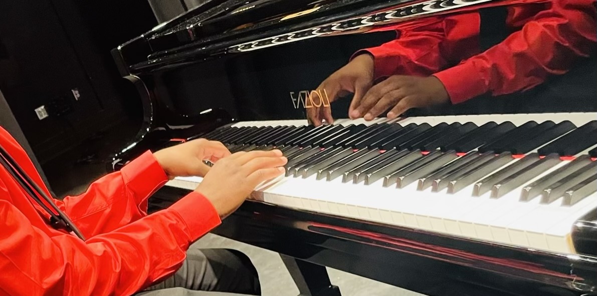 Person in red shirt playing piano, hands on keys, reflection visible on glossy surface, arm and hand details visible.