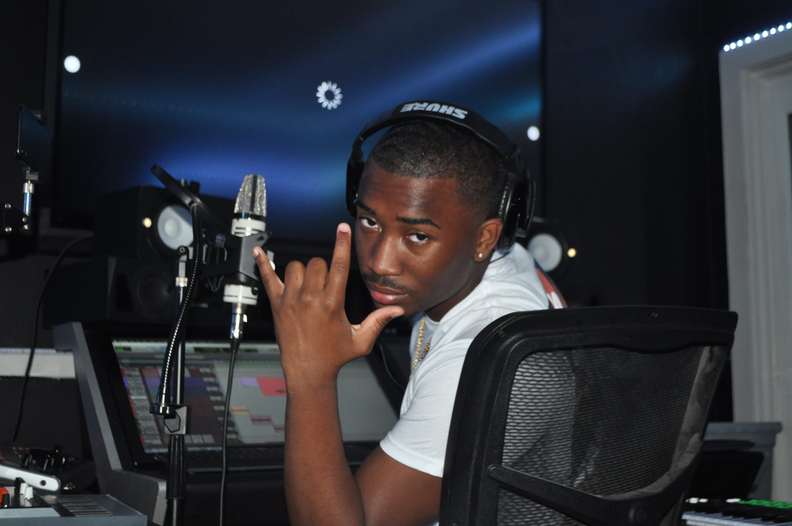 Young man wearing headphones in a recording studio, sitting at a mixing console, looking at the camera with a serious expression.