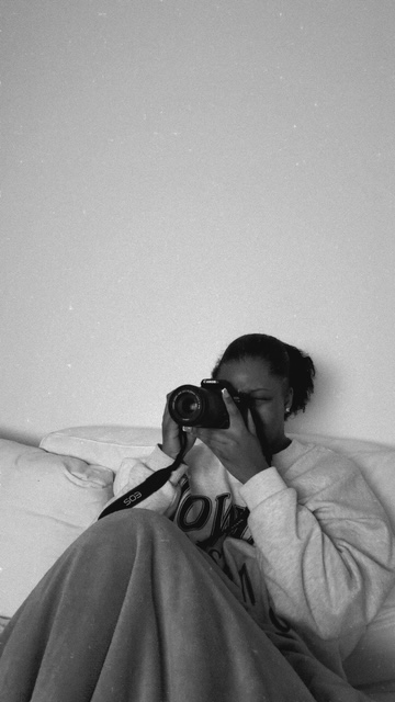 Person sitting on bed taking a photo with a camera, wearing a hoodie, in a room with plain wall
