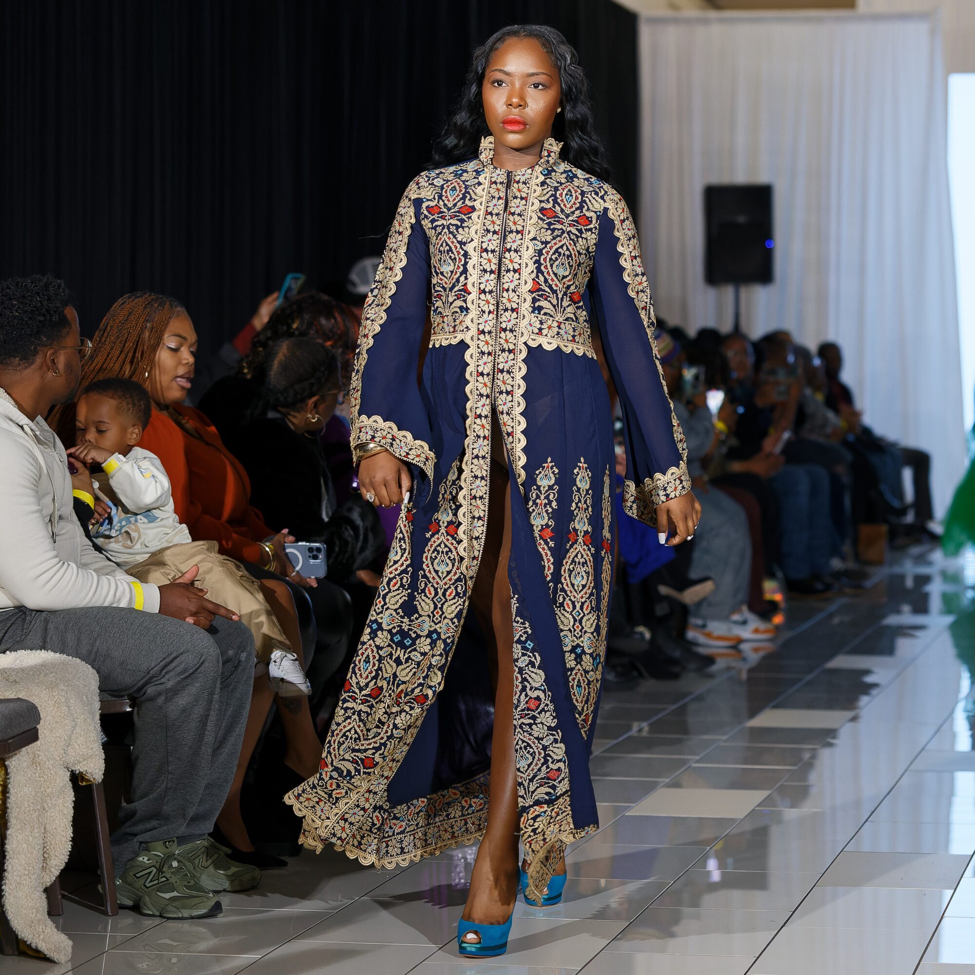 Model walking on runway in a long, ornate blue and gold embroidered gown with a high slit, audience seated on sides.