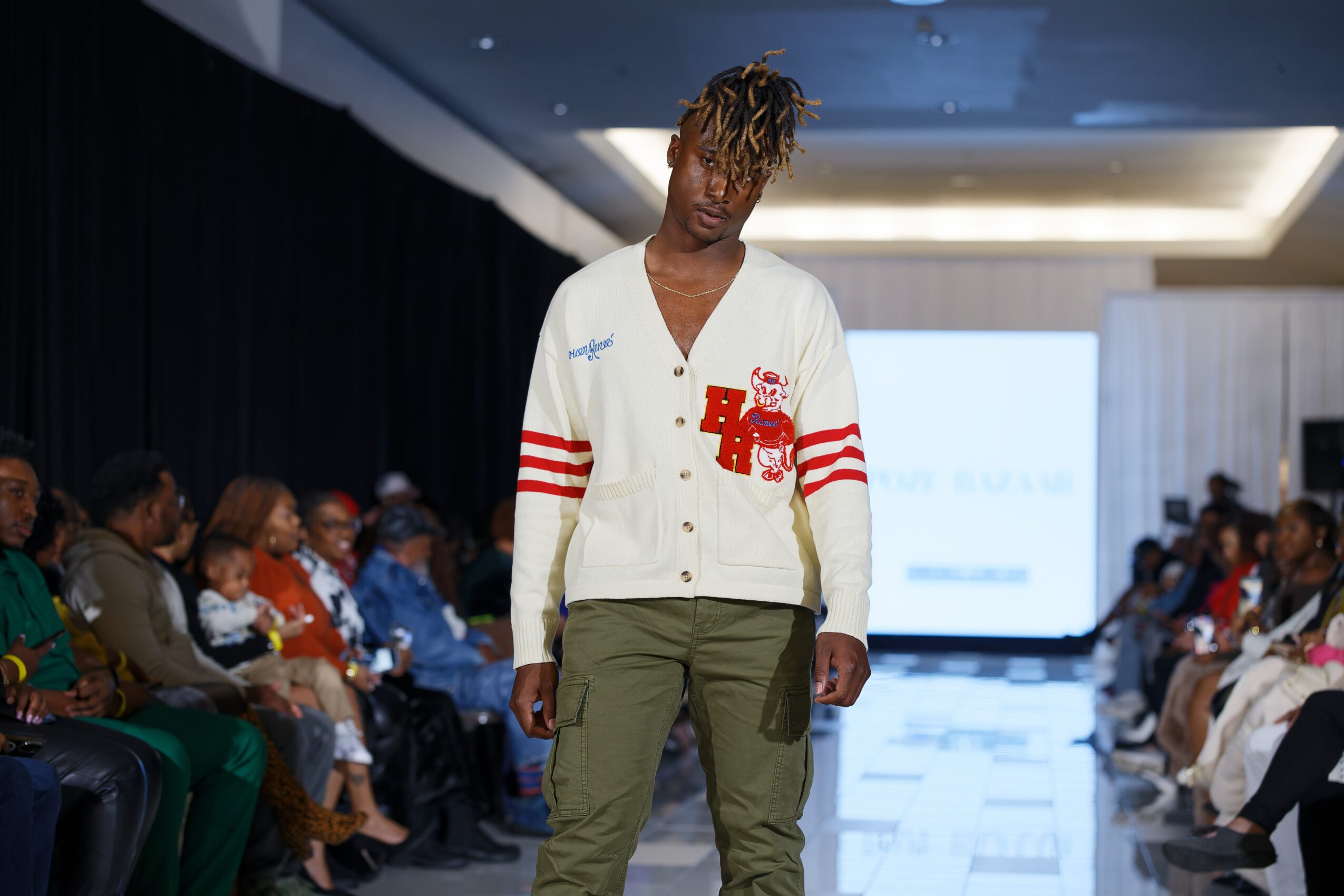 Person walking on runway during fashion show, audience seated on both sides, large screen in background, indoor setting