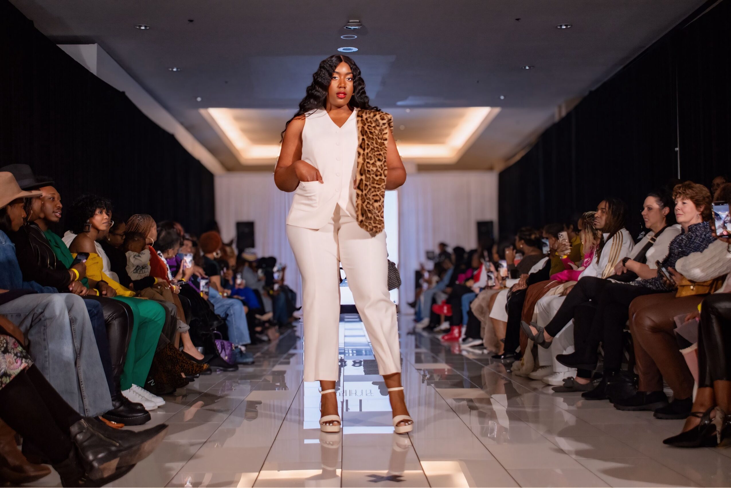 Model walking on runway with audience on both sides, wearing white outfit and leopard print scarf, in a fashion show setting.