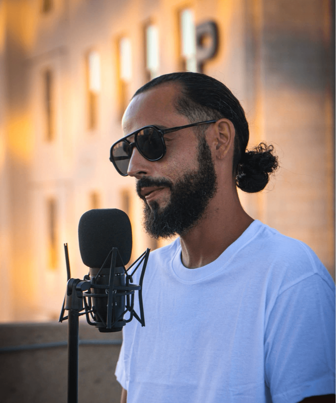 Man with sunglasses and beard speaking into a microphone outdoors during evening, blurred building background.