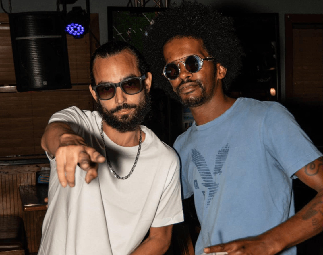 Two men with sunglasses pose indoors, one with a beard and chain, the other with curly hair and a blue shirt.
